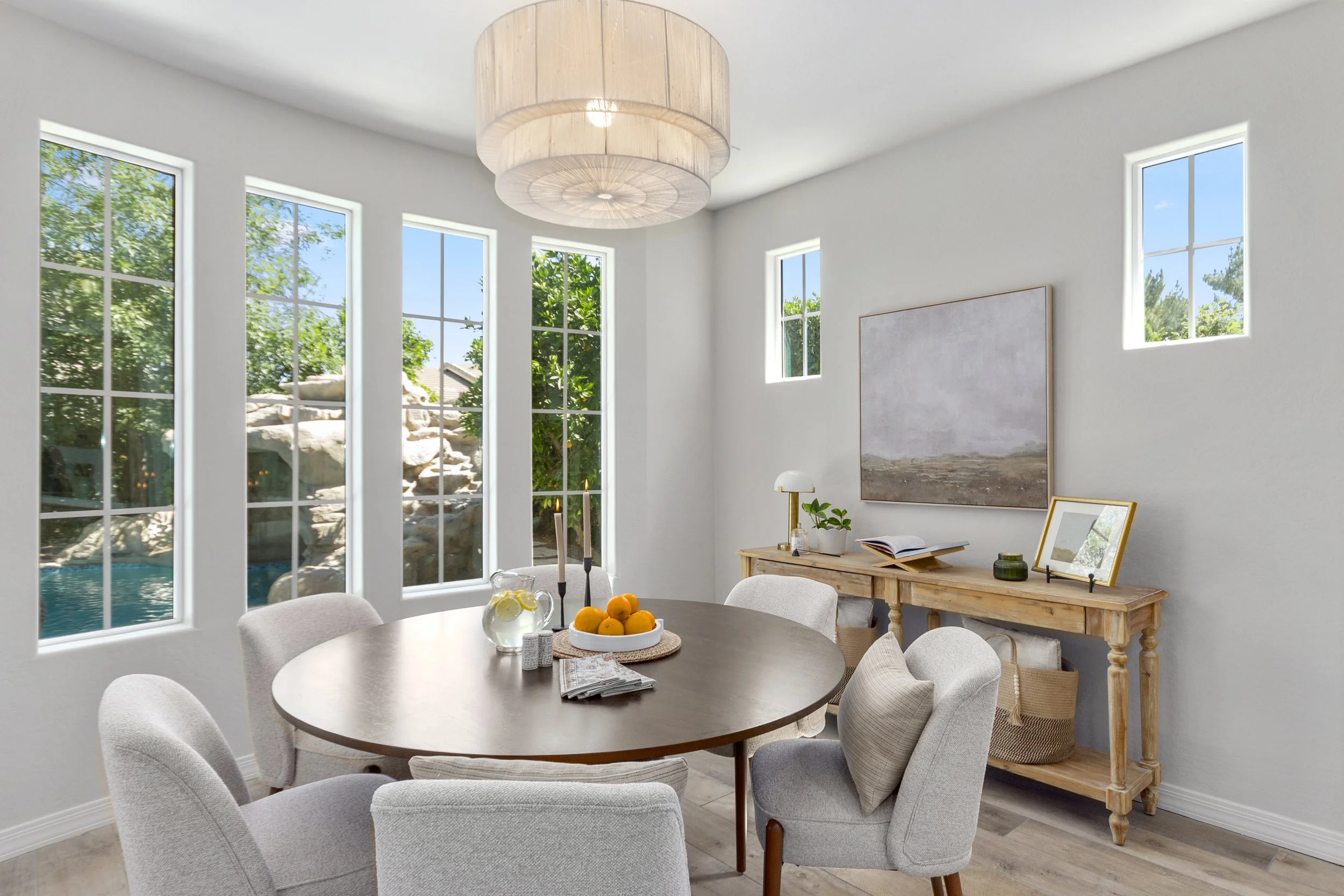 A bright dining room with white walls and large windows revealing a garden outside. A round wooden table surrounded by cushioned chairs holds a bowl of oranges and a glass pitcher of water with lemon slices. A wooden sideboard against the wall displa