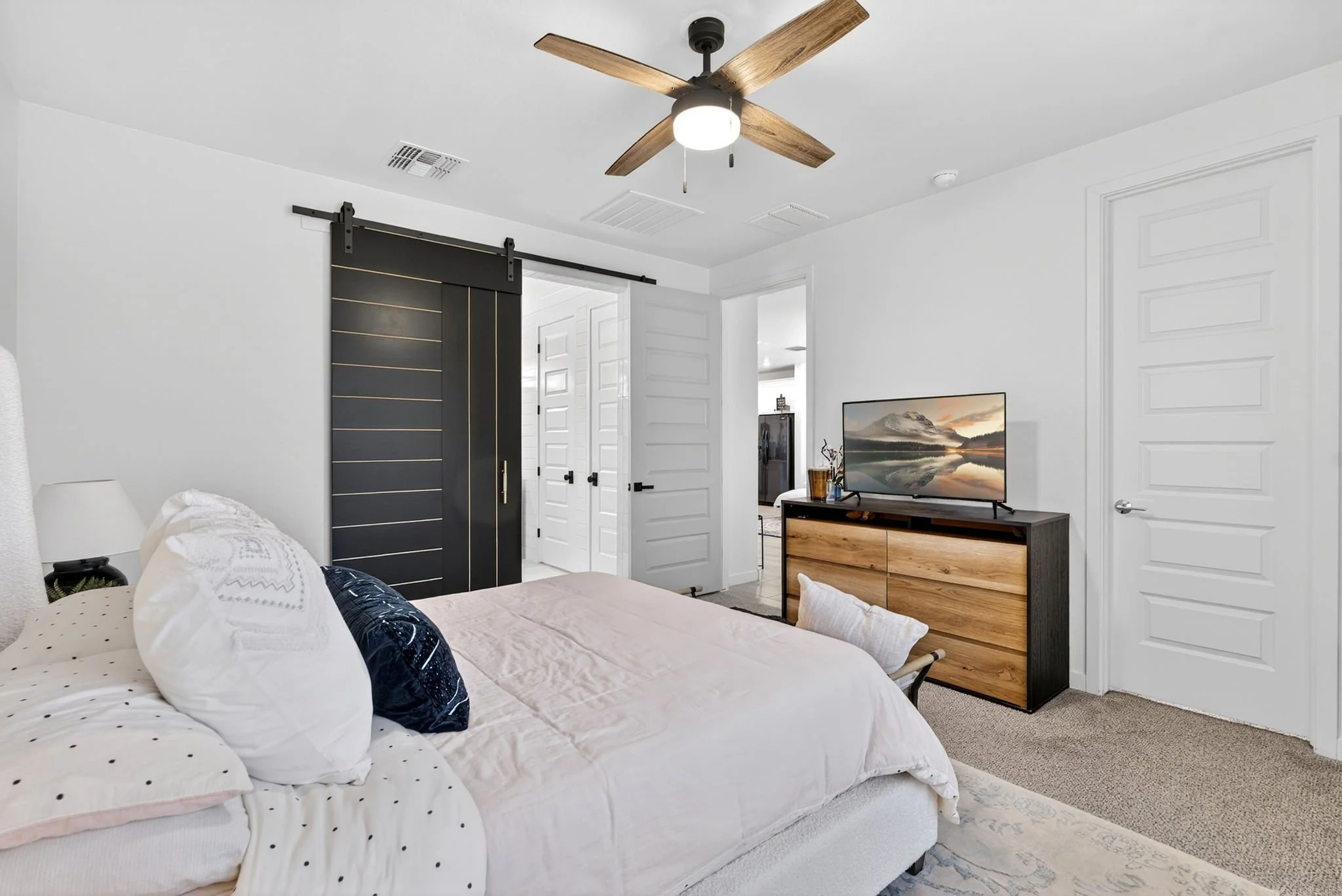 A bedroom with a white bed, decorative pillows, a wooden dresser, a flat-screen TV, and white walls with paneled doors. A ceiling fan with wooden blades is on the ceiling.