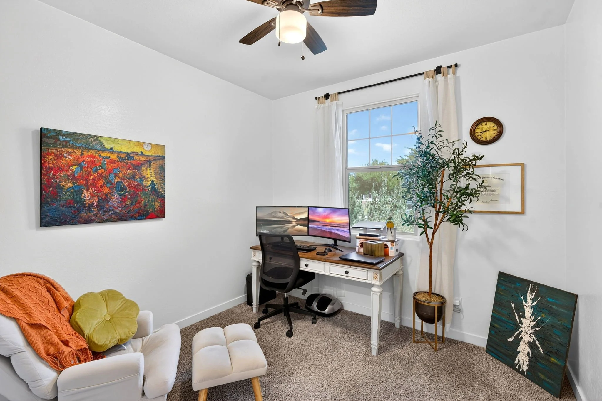 A cozy home office with white walls and a window with white curtains. The room features a white desk with dual monitors, a black office chair, and decorative artwork including colorful paintings hanging on the walls and leaning on the floor. A ceilin