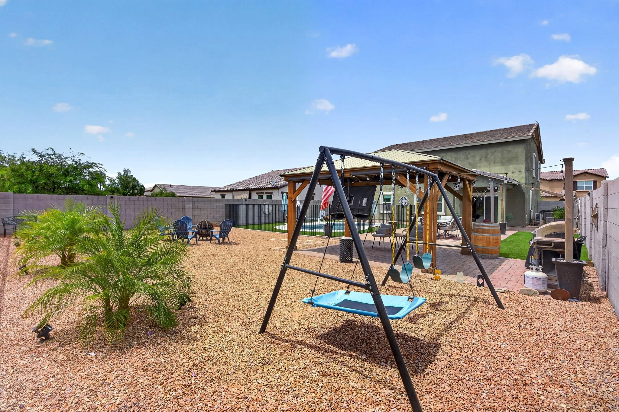 Backyard with a playground swing set, surrounded by gravel, patio furniture, and landscaping with a green shrub, with houses visible in the background under a blue sky with scattered clouds.