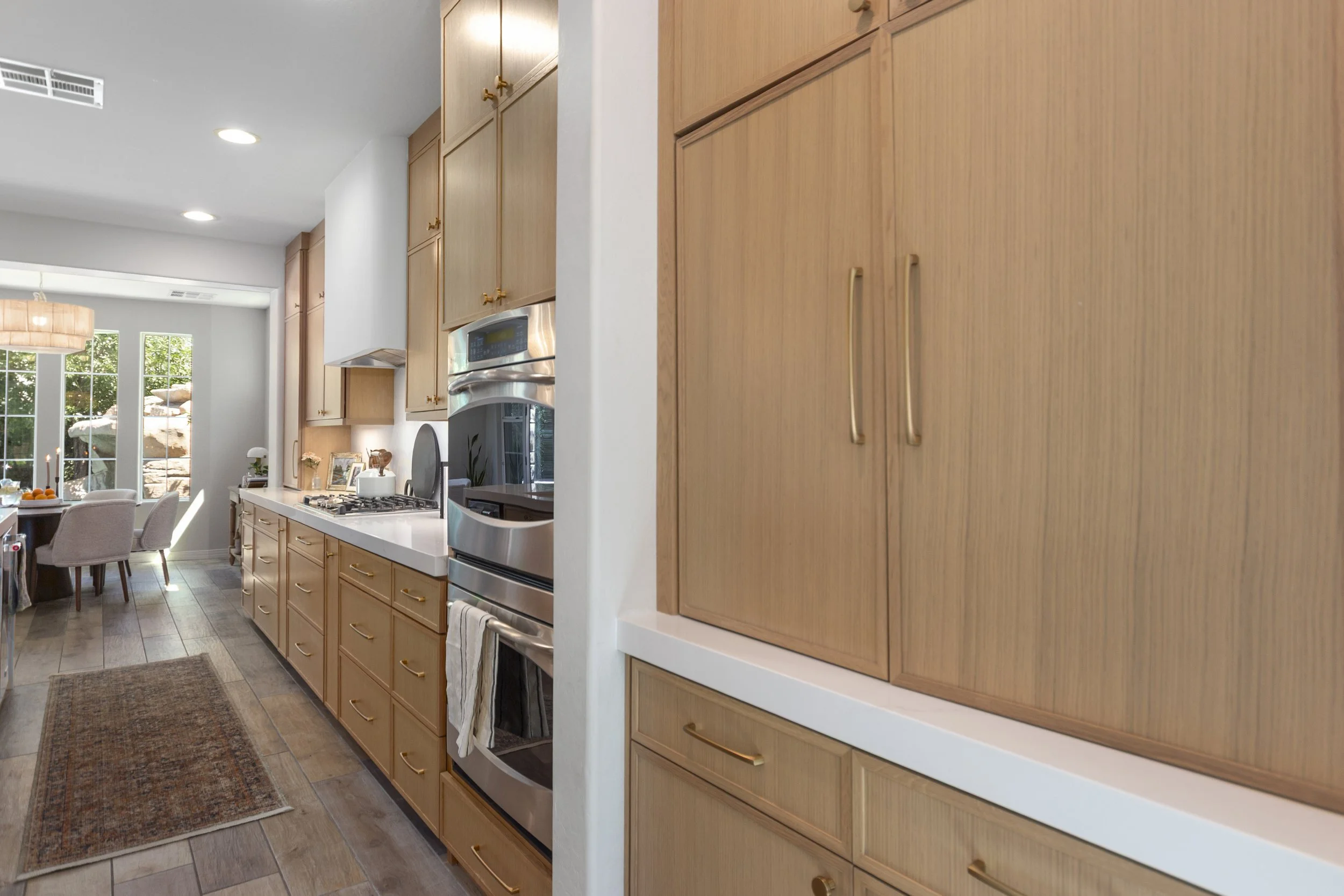 Kitchen with wooden cabinets, white walls, a stainless steel oven, and a dining area with beige chairs near large windows showing green outdoor scenery.