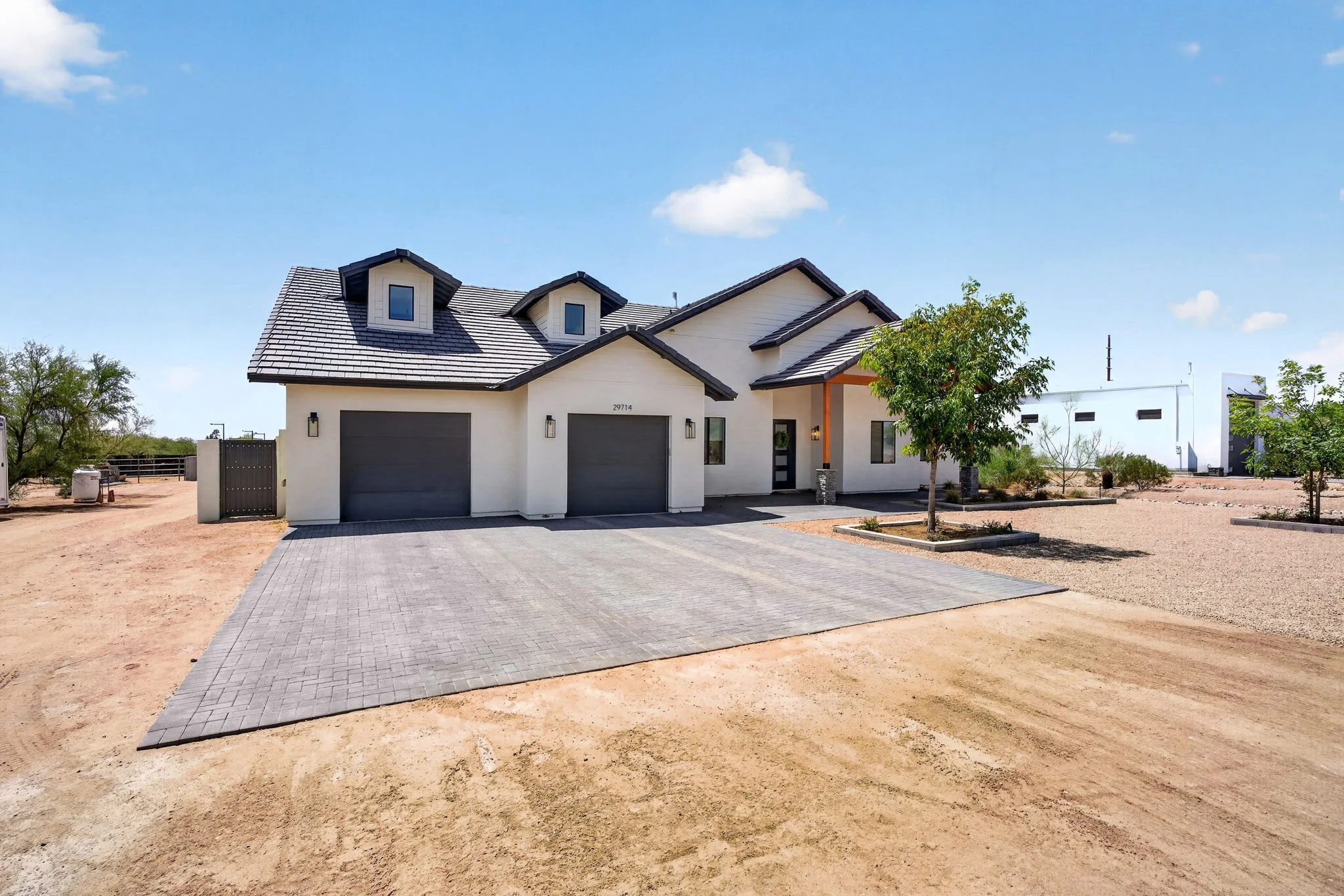 A modern two-story house with a gray garage door, a small front porch, and a landscaped yard with trees, situated on a paved driveway and surrounding dirt area under a blue sky.