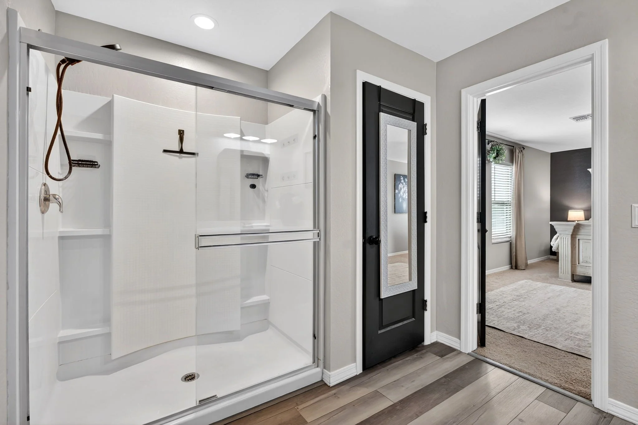 A bathroom with a glass shower enclosure, beige walls, a black door with a mirror, and a view into a bedroom with a window, curtains, and a bed.