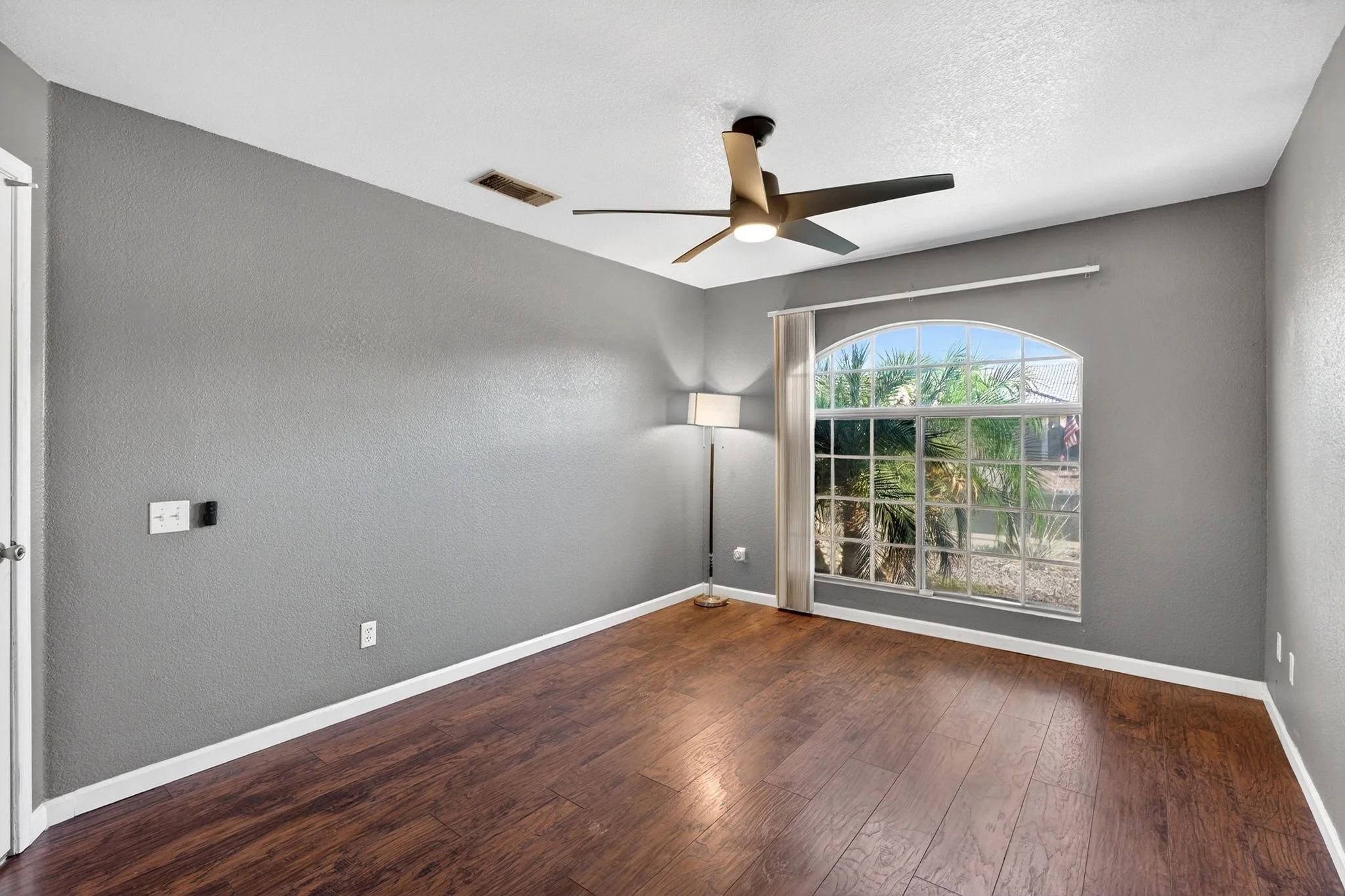 Empty living room with light gray walls, large arched window with white blinds, dark wood flooring, a standing lamp, and a ceiling fan with integrated light.