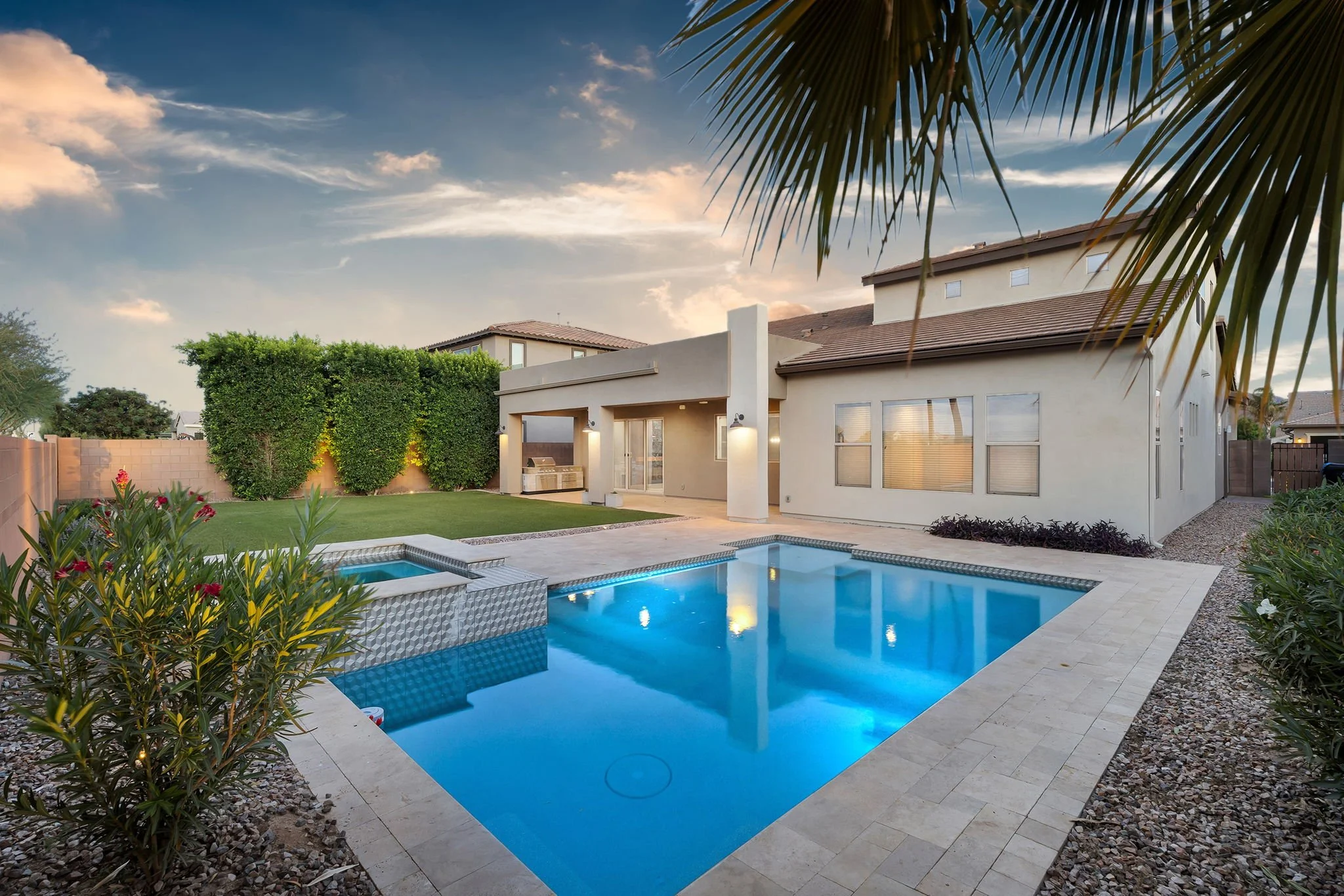 Modern backyard with a swimming pool, surrounded by a stone deck, lush bushes, and plants, with a white house in the background, under a partly cloudy sky.
