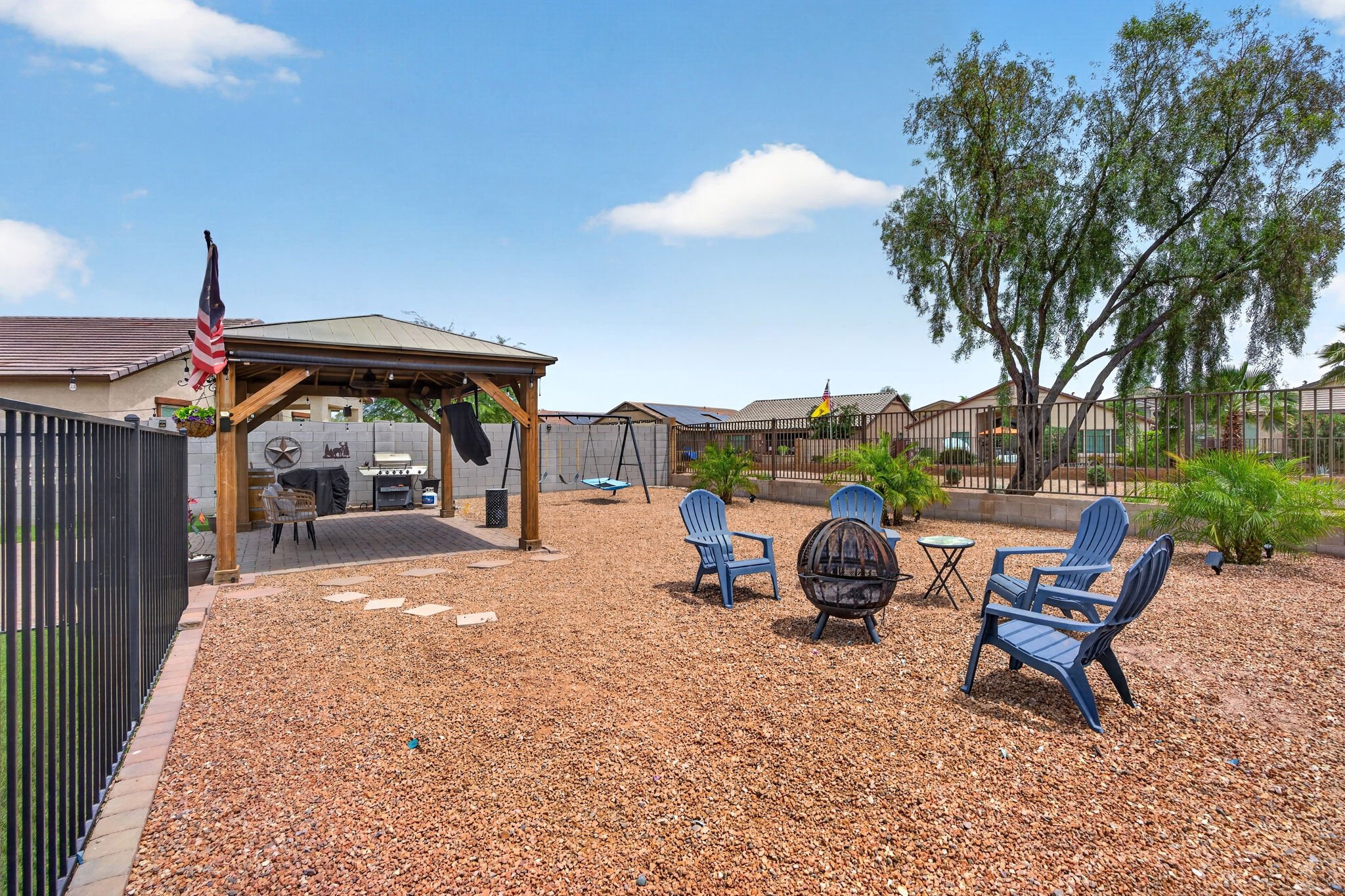 Backyard with gravel ground, blue patio chairs, a fire pit, trees, and a white fence. There is a covered area with outdoor furniture, a grill, and a swing set. Flags are visible in neighboring yards under a partly cloudy sky.