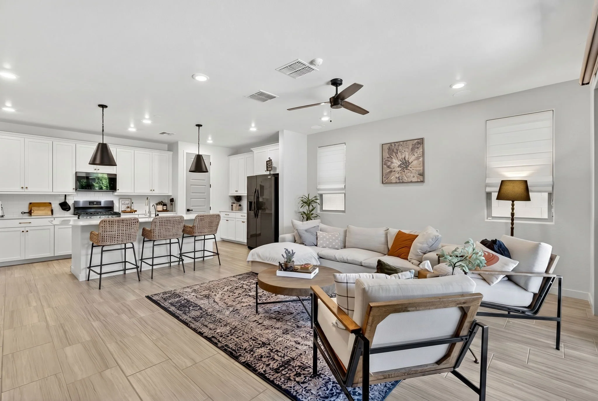 Open-concept living room and kitchen area with white cabinets, black appliances, pendant lights, and wicker bar stools at a white kitchen island, a ceiling fan, framed artwork, and a beige sofa with pillows.
