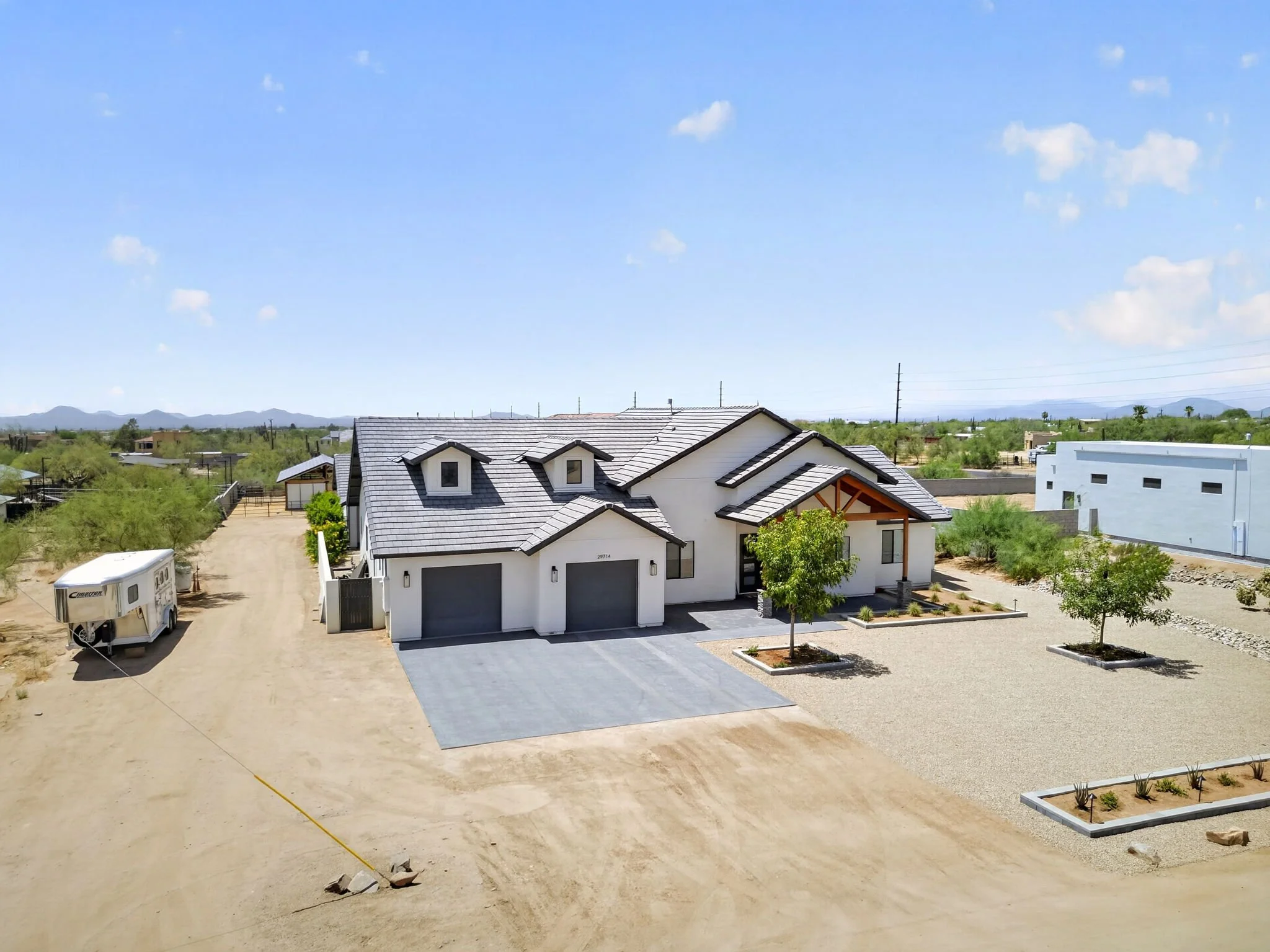 A modern white house with a gray roof, a driveway, and a small front yard with plants and trees, in a desert landscape with mountains in the background under a blue sky with scattered clouds.