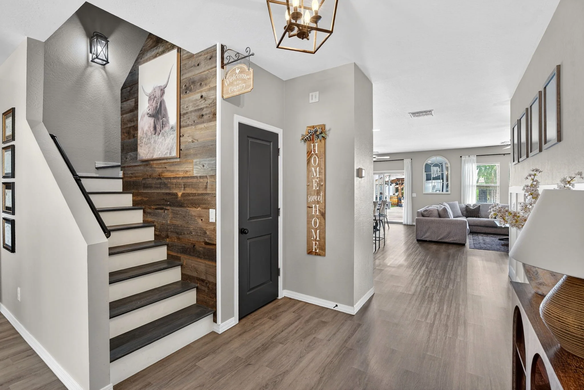 Interior view of a living room and entryway with stairs. The stairs have dark wood steps opposite white risers. The wall near the stairs has framed pictures. The entryway has a dark door, and decorative signs including one that says 'Welcome in our C