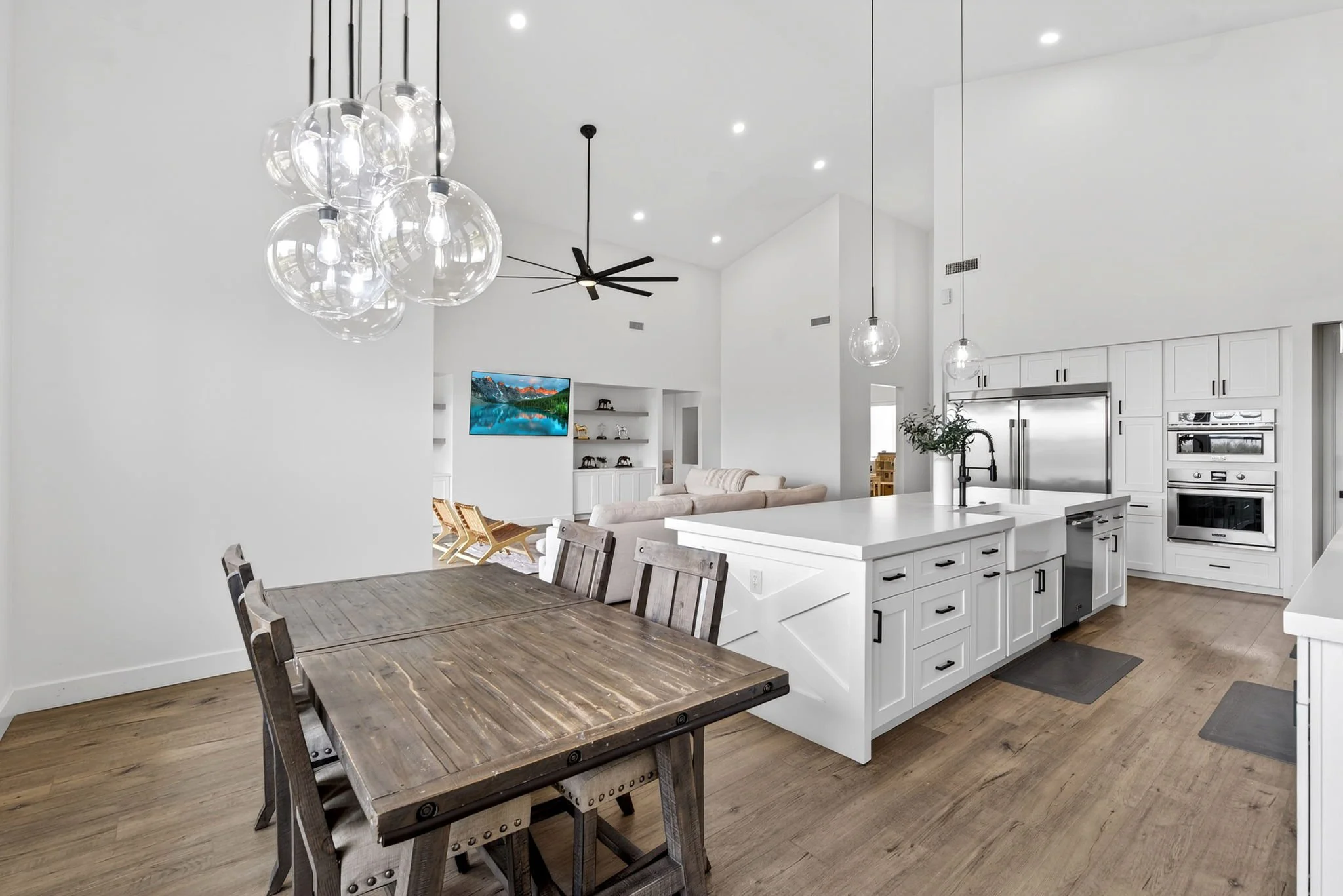 Open-concept kitchen and dining area with white walls, wooden flooring, a rustic wooden dining table with matching chairs, a large kitchen island with a white countertop, modern light fixtures, a ceiling fan, built-in appliances, and a flat-screen TV