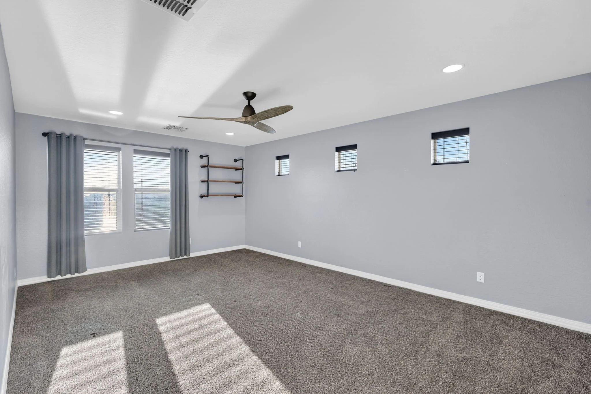 Empty room with gray walls, gray curtains, three small high windows with blinds, a larger window with blinds, a ceiling fan with wooden blades, ceiling lights, gray carpet, and a wall-mounted shelf.