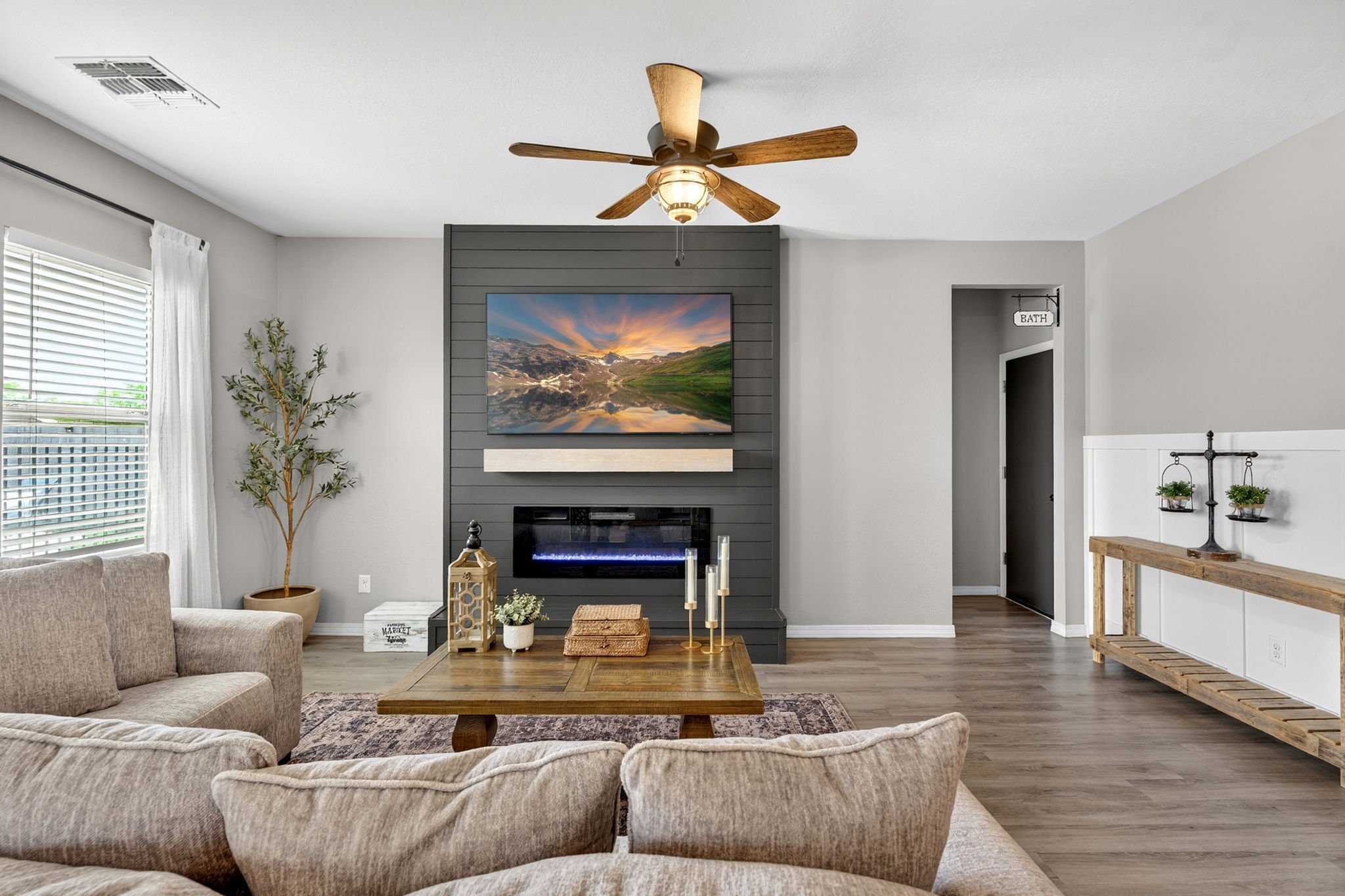 Living room with beige sofas, a wooden coffee table, a fireplace with a television above, a potted plant, and decorative items. Large window with white curtains, ceiling fan, and a hallway with a bath sign visible.
