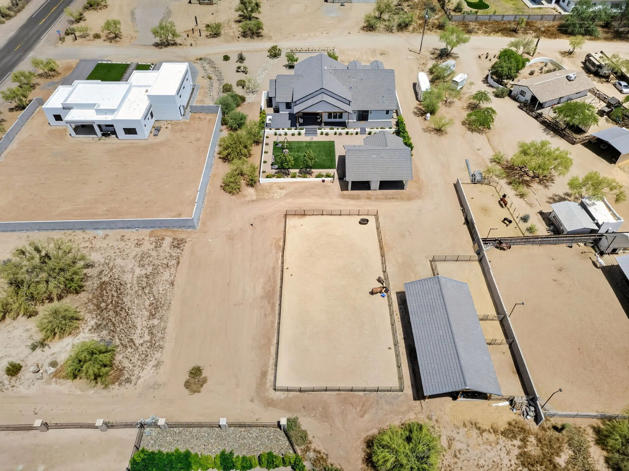 Aerial view of a residential area featuring multiple houses, some with modern designs and others traditional, with yards and trees, dirt roads, and a fenced-in area with horses.