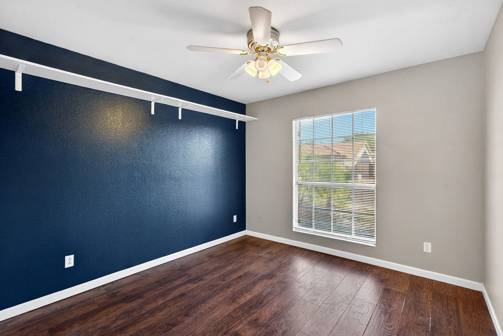 Empty room with a navy accent wall, white ceiling fan with lights, large window with blinds, hardwood flooring, and white baseboards.