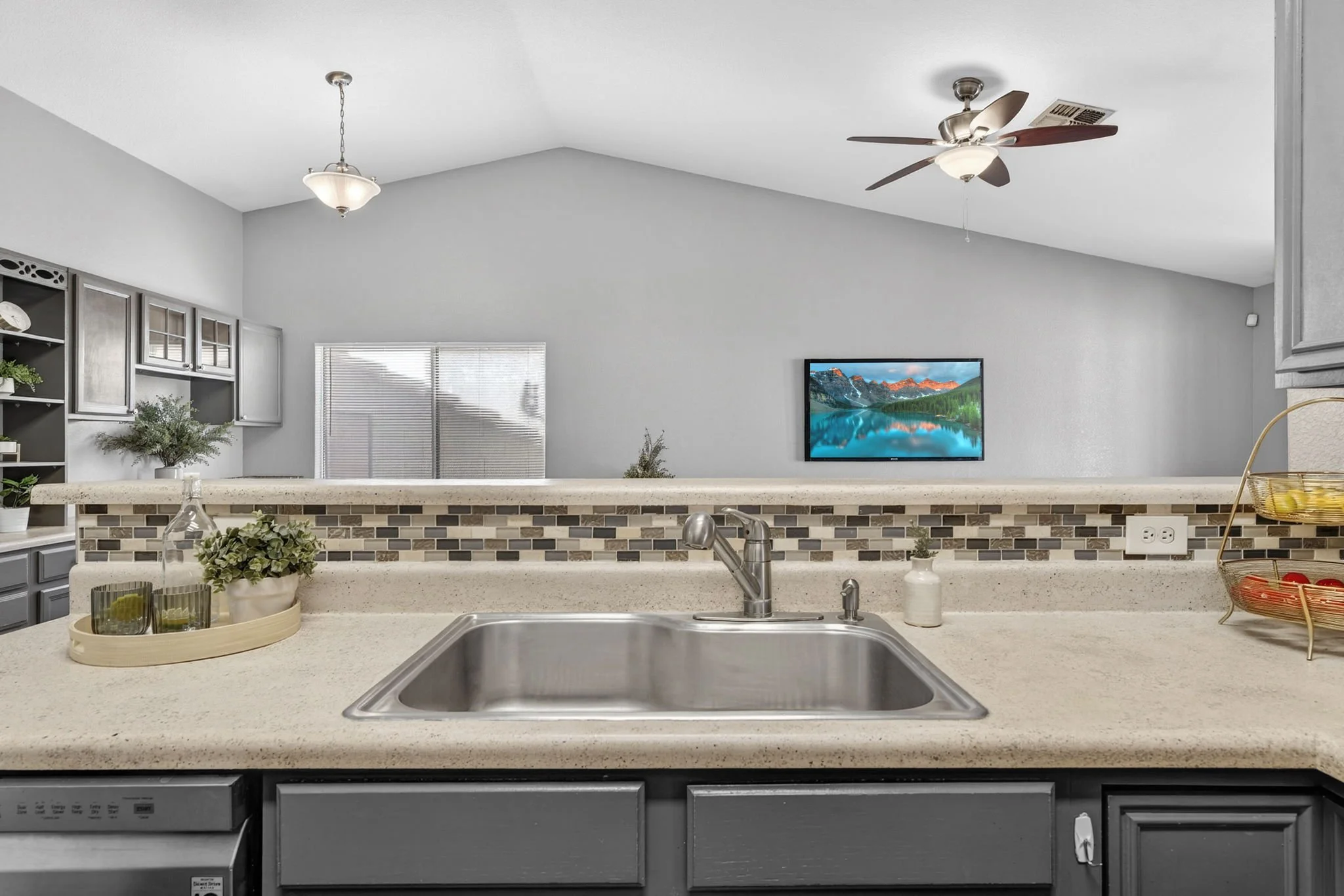 View of a kitchen with a beige countertop, sink, and black cabinets. There are decorative items, plants, and glassware on the counter. In the background, there is a gray wall with a mounted TV displaying a mountain lake scene, overhead lights, and a 