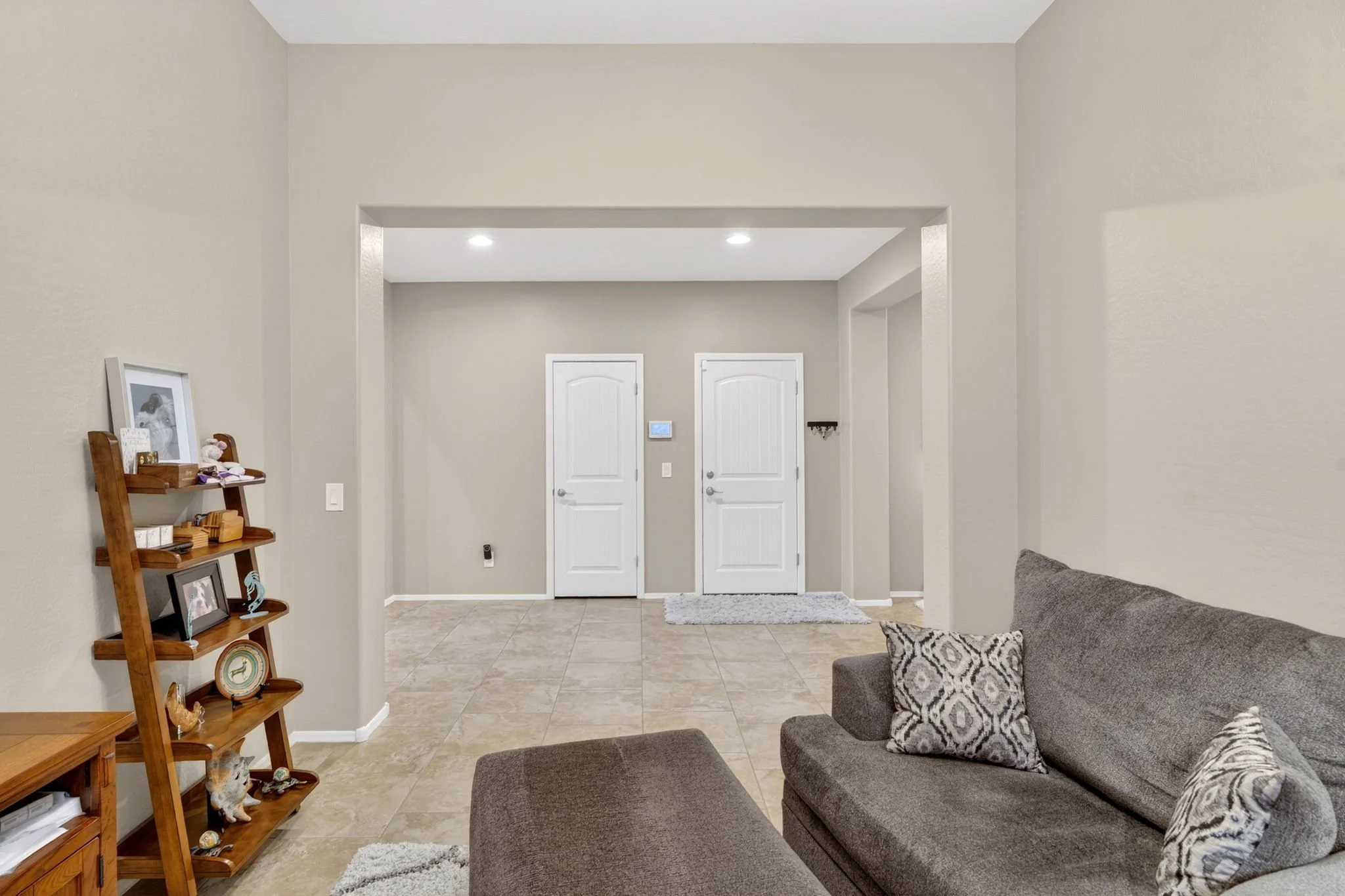 Living room with beige walls, tile floor, gray couch with patterned pillows, and a wooden ladder shelf with photos and decorations. In the background, there are two white doors and a small ottoman or rug near the entrance.
