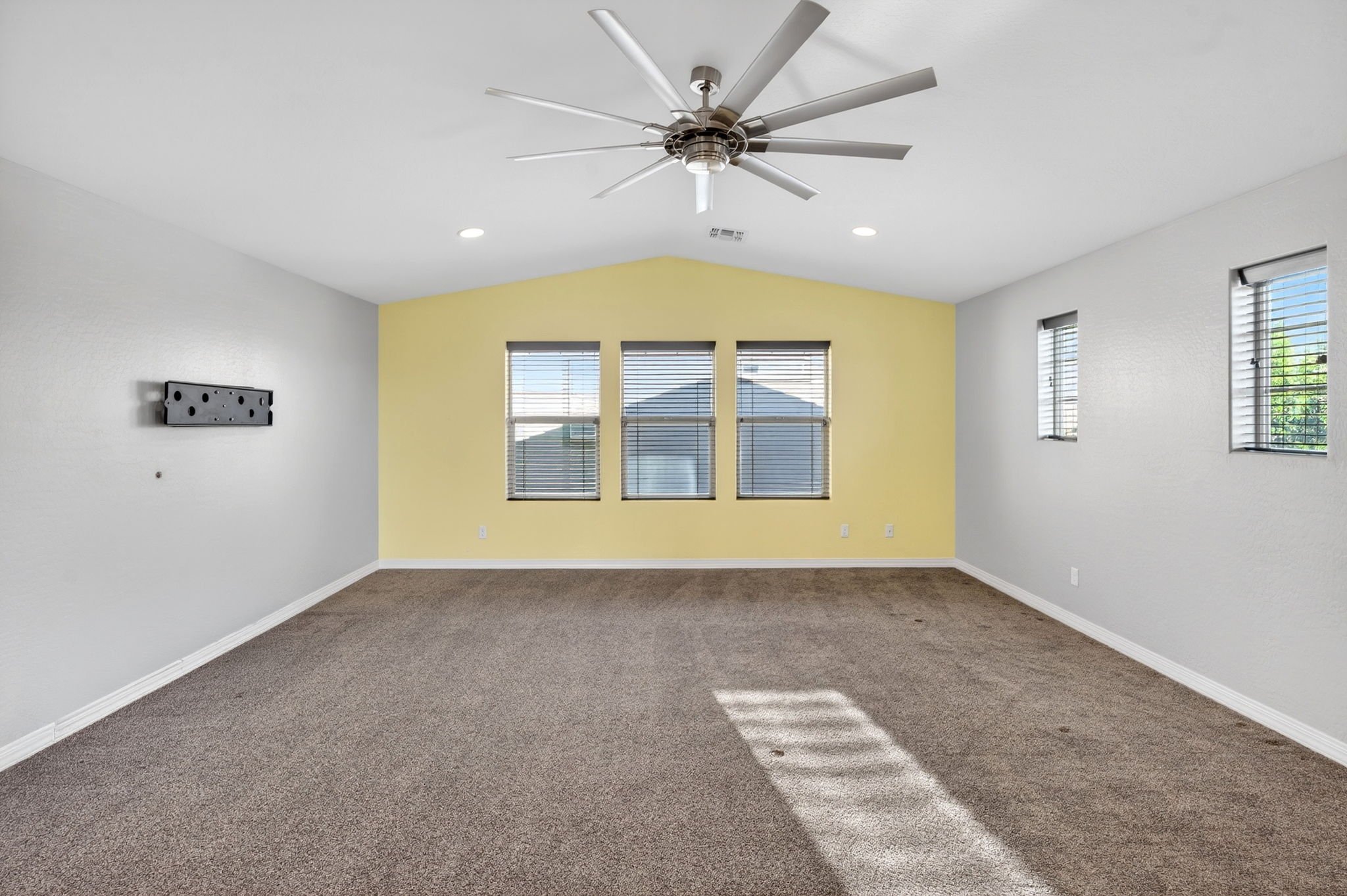 Empty living room with three windows, light yellow accent wall, gray walls, ceiling fan, and carpeted floor.