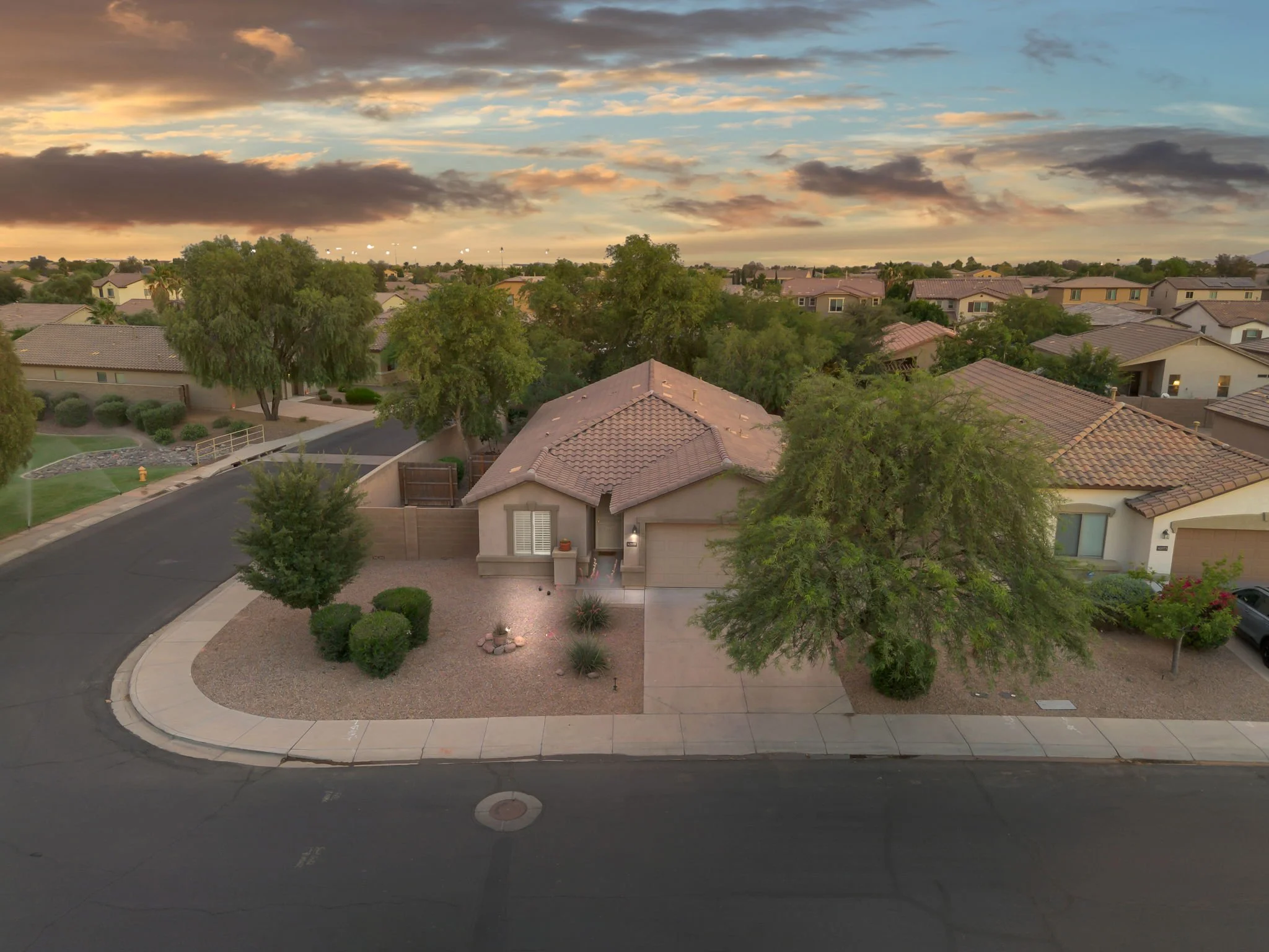 A suburban neighborhood street at dusk with single-family homes, green trees, and a cloudy sky.