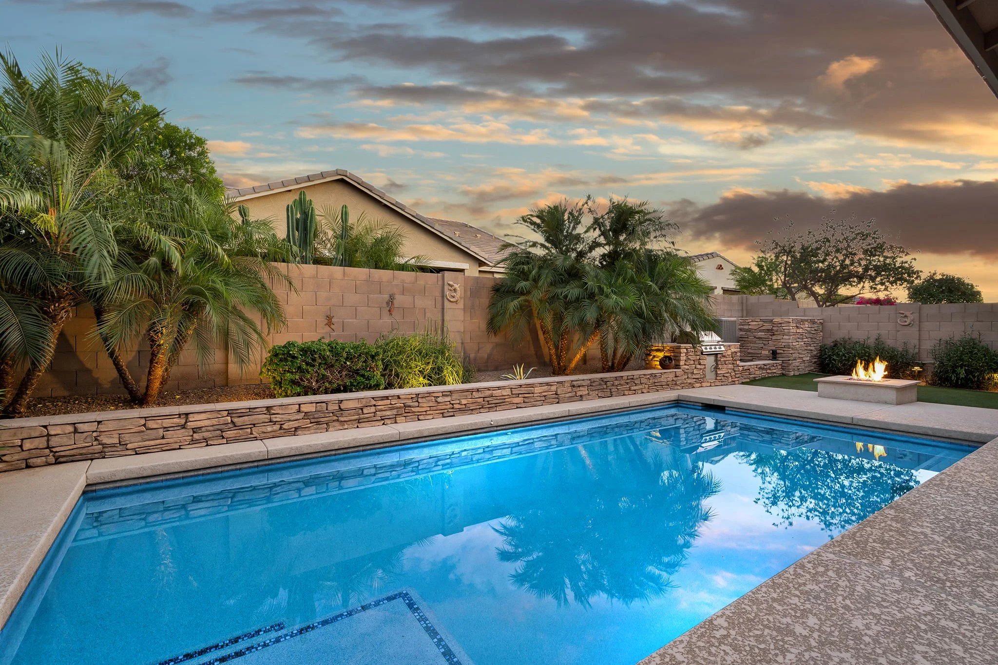 Backyard with a swimming pool, stone wall, lush green plants, trees, and a fire feature, under a partly cloudy sky at sunset.