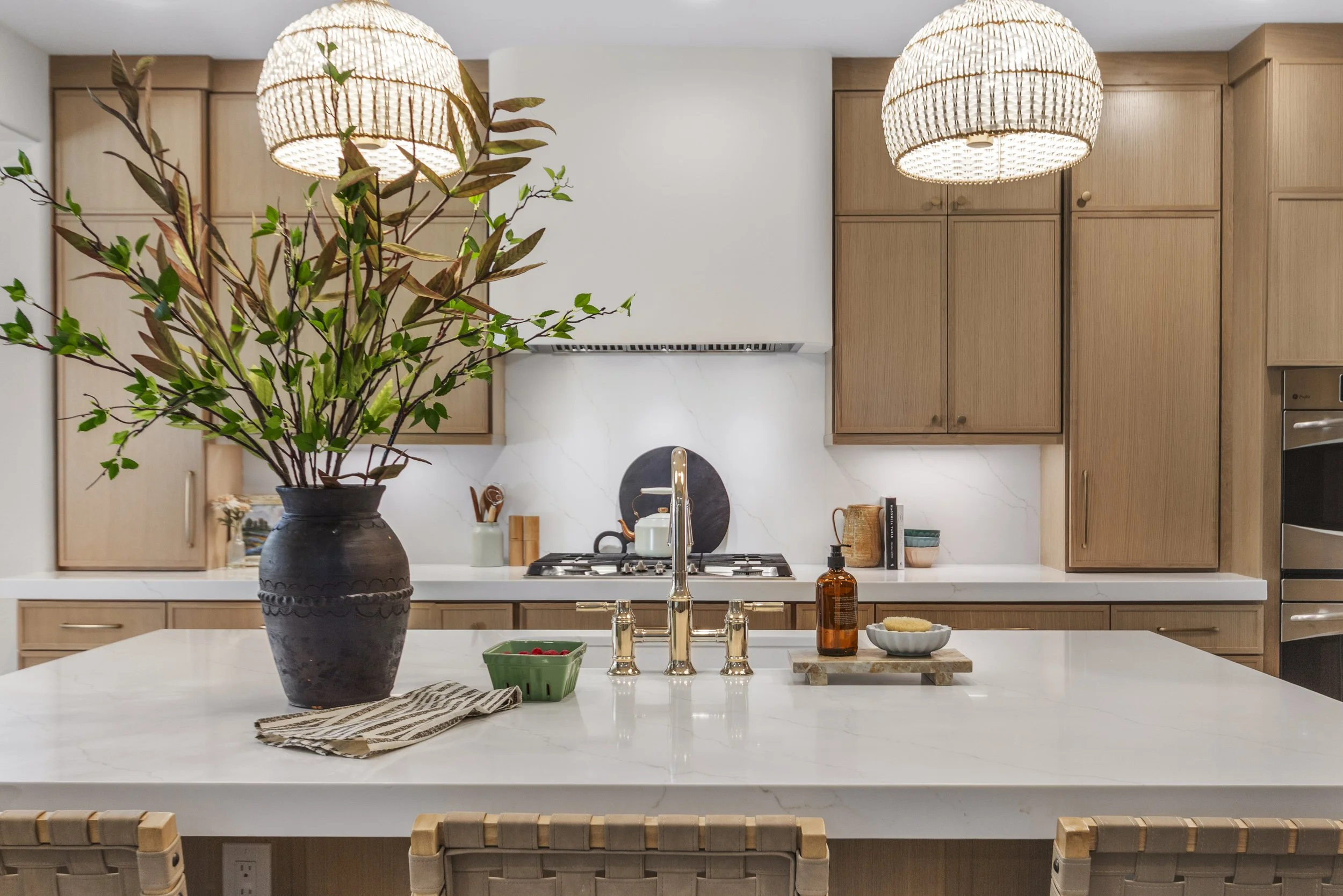 A modern kitchen with a white marble island, a large potted plant, and wooden cabinetry. There are two woven pendant lights hanging from the ceiling and various kitchen items on the counter.