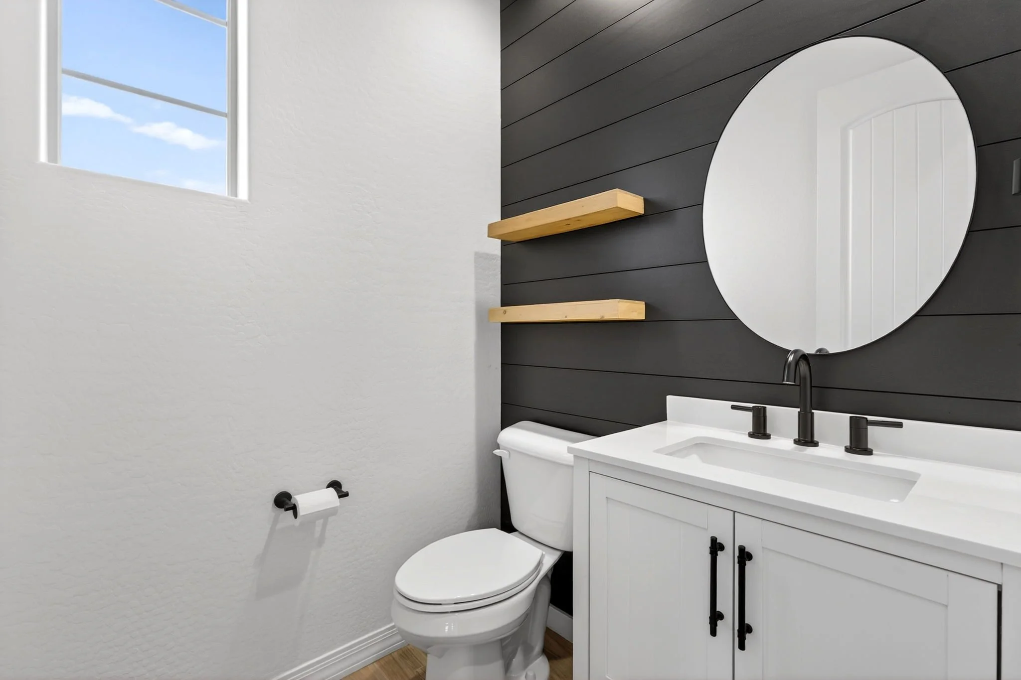 A modern bathroom with a white toilet, a white vanity with a black faucet, a round mirror, and two floating wooden shelves against a black shiplap wall. There's a window with a view of a blue sky with clouds.
