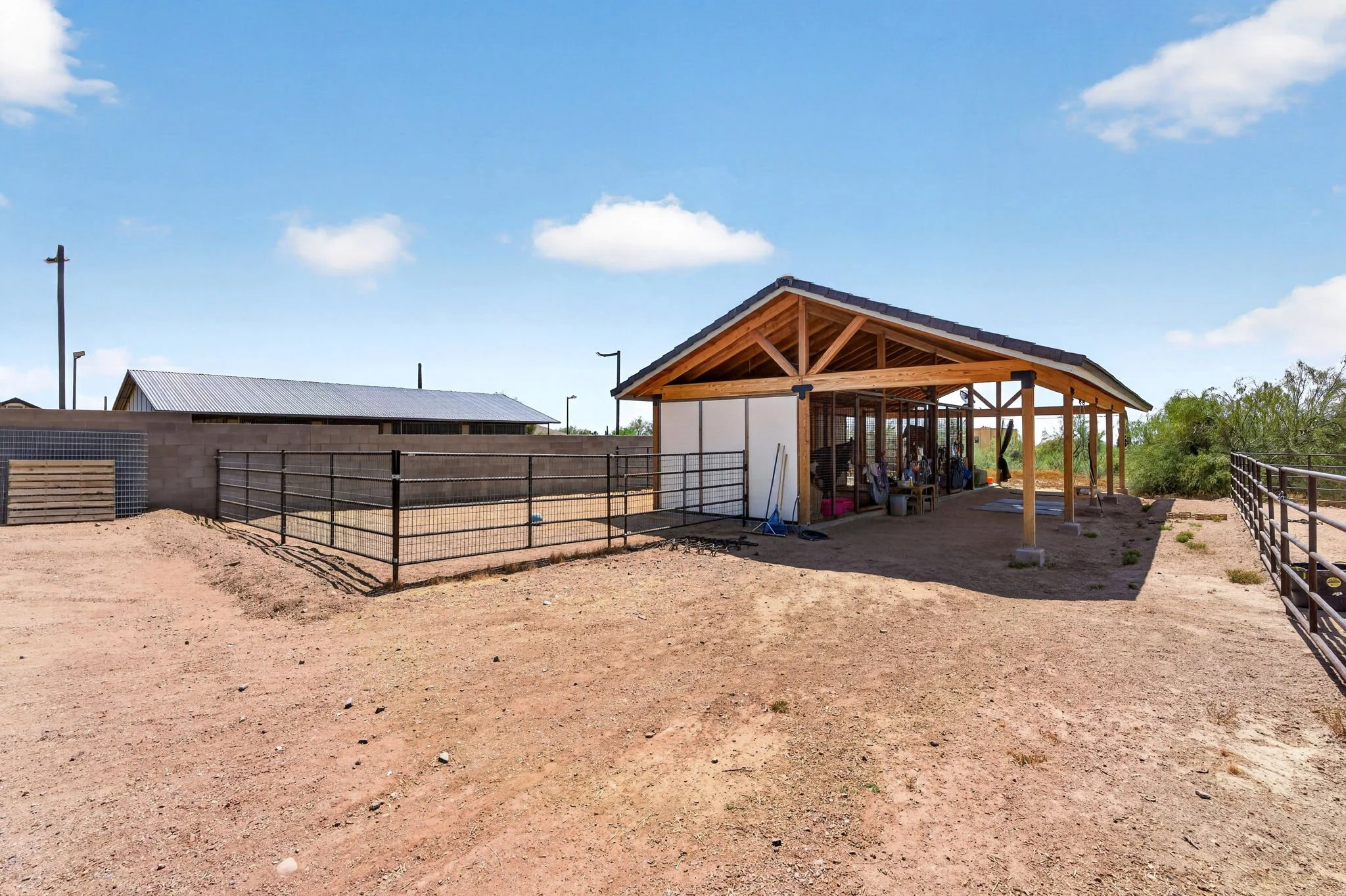 A partly constructed building with a wooden roof and open sides, surrounded by a black metal fence on a dirt lot with a clear blue sky and some clouds.
