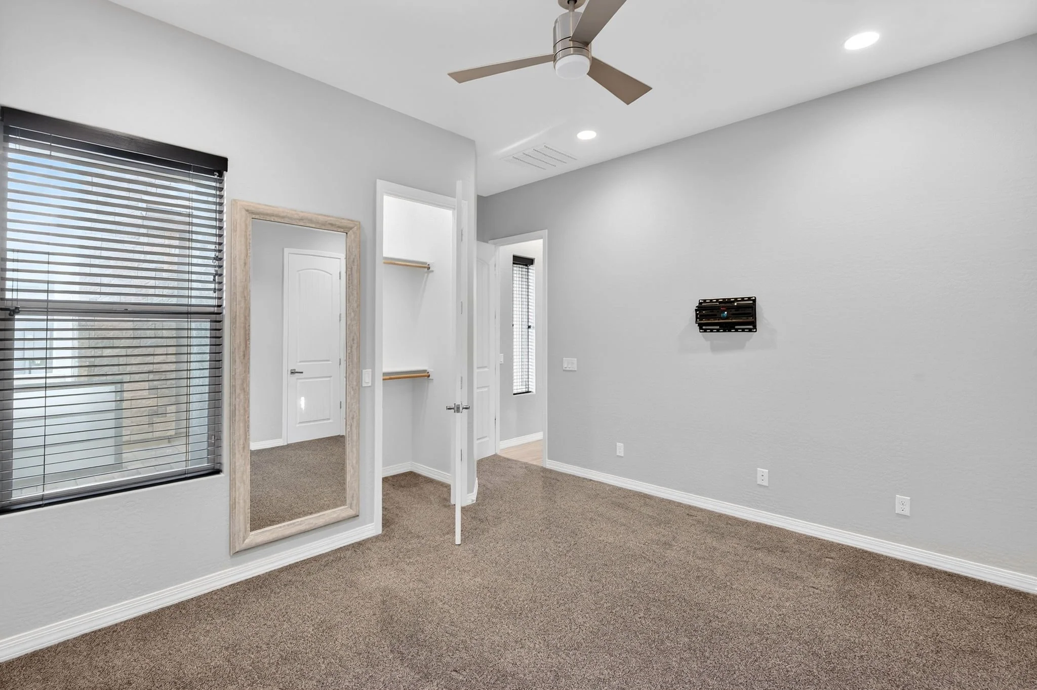 Interior room with light gray walls, beige carpet, a standing mirror with a wood frame, and windows with black blinds. There is a small wall-mounted black shelf on the right wall and ceiling lights, including a fan with lights.