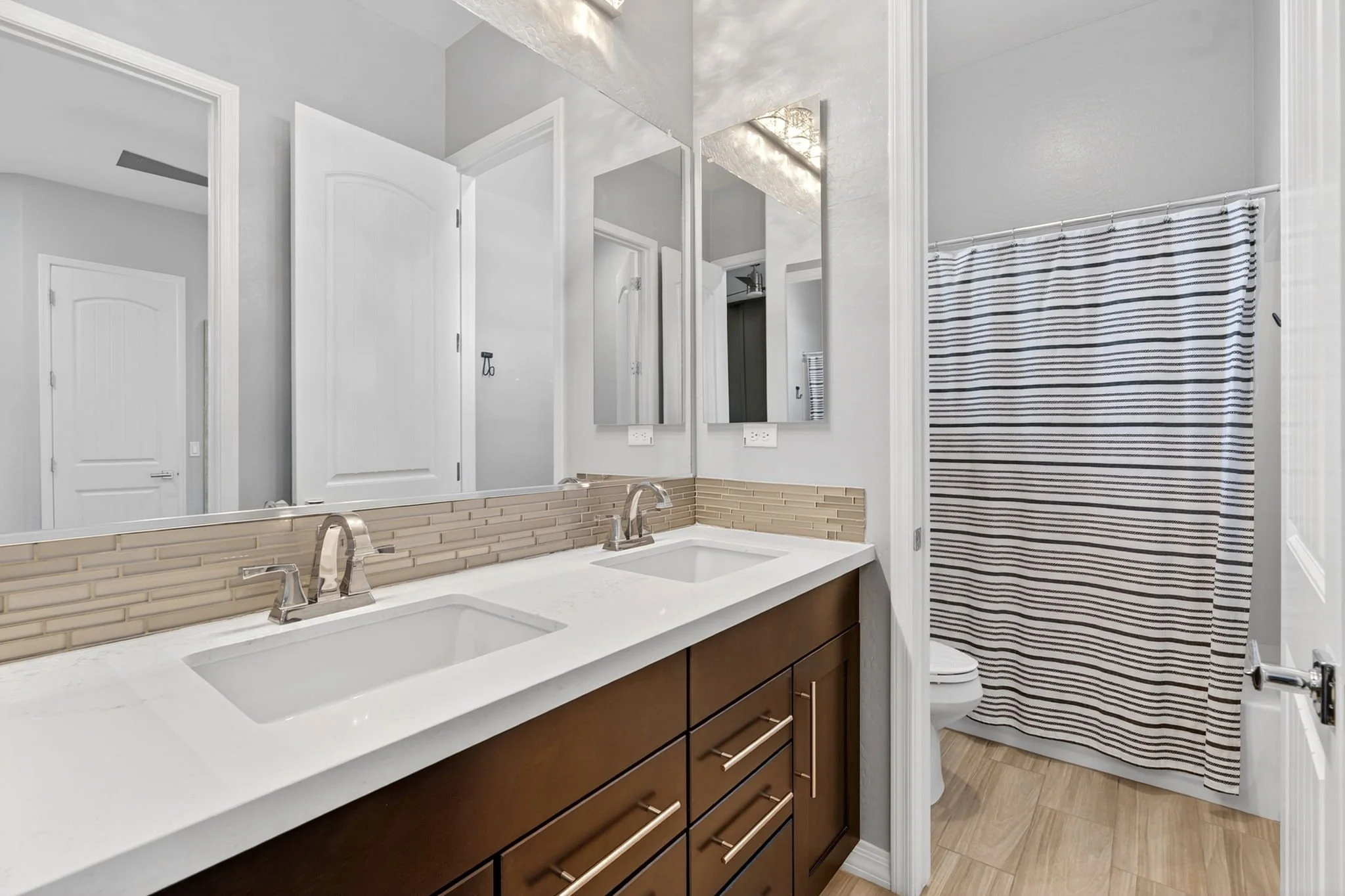 Modern bathroom with double sink vanity, large mirror, striped shower curtain, neutral wall colors, and wood flooring.