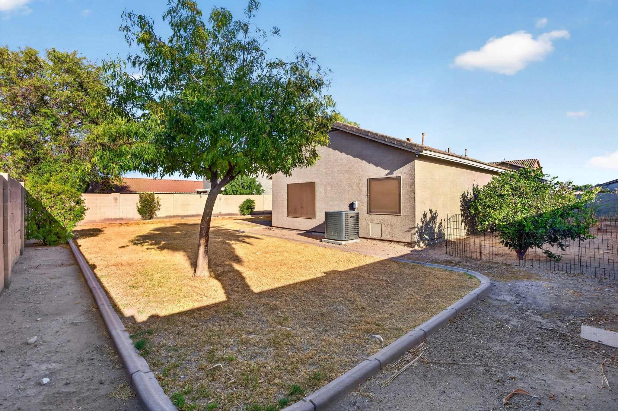 A backyard with a tree, dry grass, a concrete path, a house wall with boarded windows, an air conditioning unit, and a small fenced area with other plantings. The sky is blue with a few clouds.