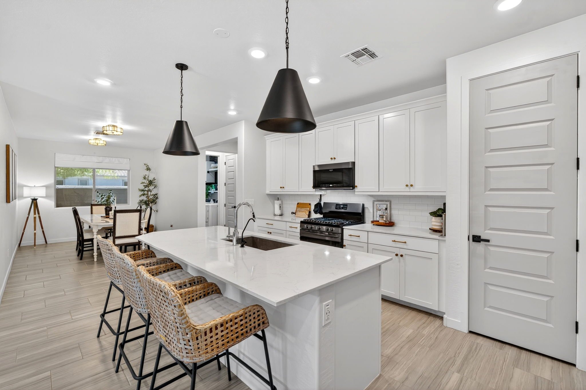 Modern kitchen with white cabinets, black pendant lights, a stove, and a dining area with wicker chairs and a table near a window.