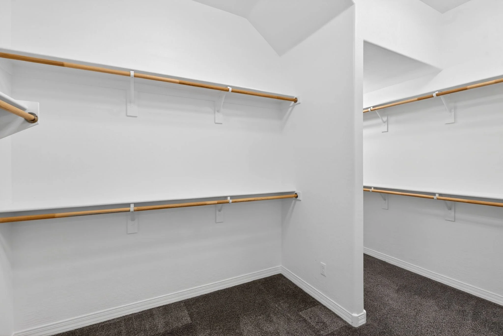 Empty walk-in closet with white walls, wooden clothing rods, and dark carpeted floor.