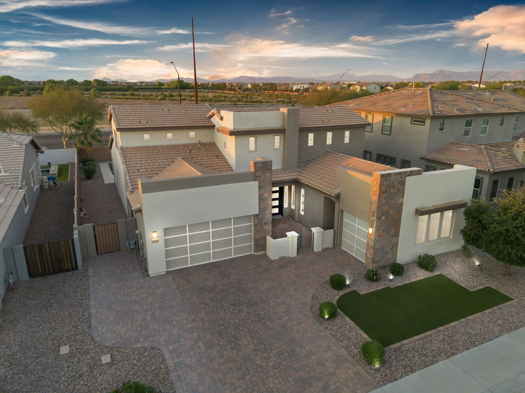 Aerial view of modern house with tiled roof, courtyard, and surrounding landscaping in a suburban neighborhood, with mountains and open land in the background during sunset.