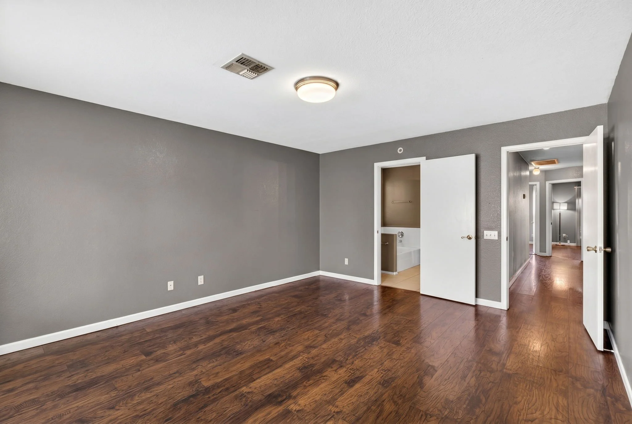 Empty room with grey walls, wooden floors, multiple doorways, a ceiling light, and a vent.