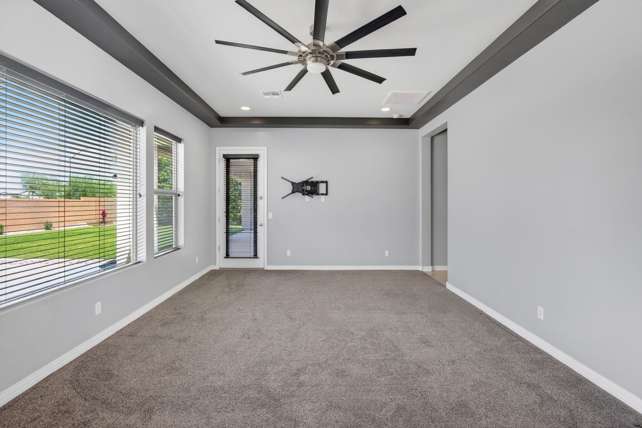 Empty living room with beige carpet, light gray walls, and large windows with blinds. Ceiling fan with multiple blades and a central light fixture.