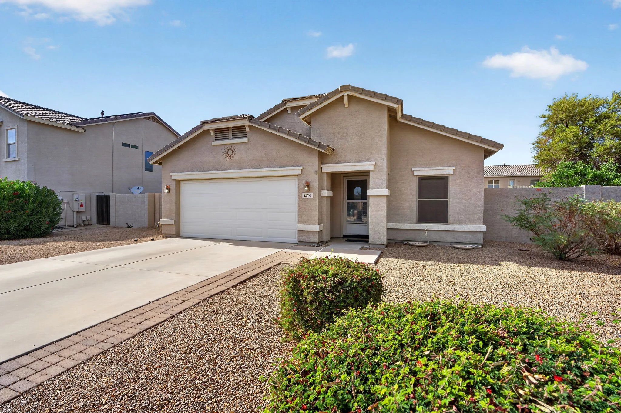 A beige two-story house with a white garage door, front door, and black window screens, along with landscaping including bushes, gravel, and a concrete driveway under a blue sky.