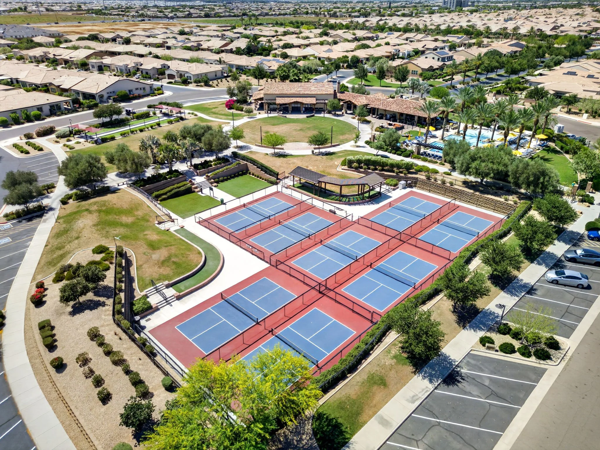 Aerial view of a residential community with houses, a park, tennis courts, a swimming pool, parking lots, and roads.