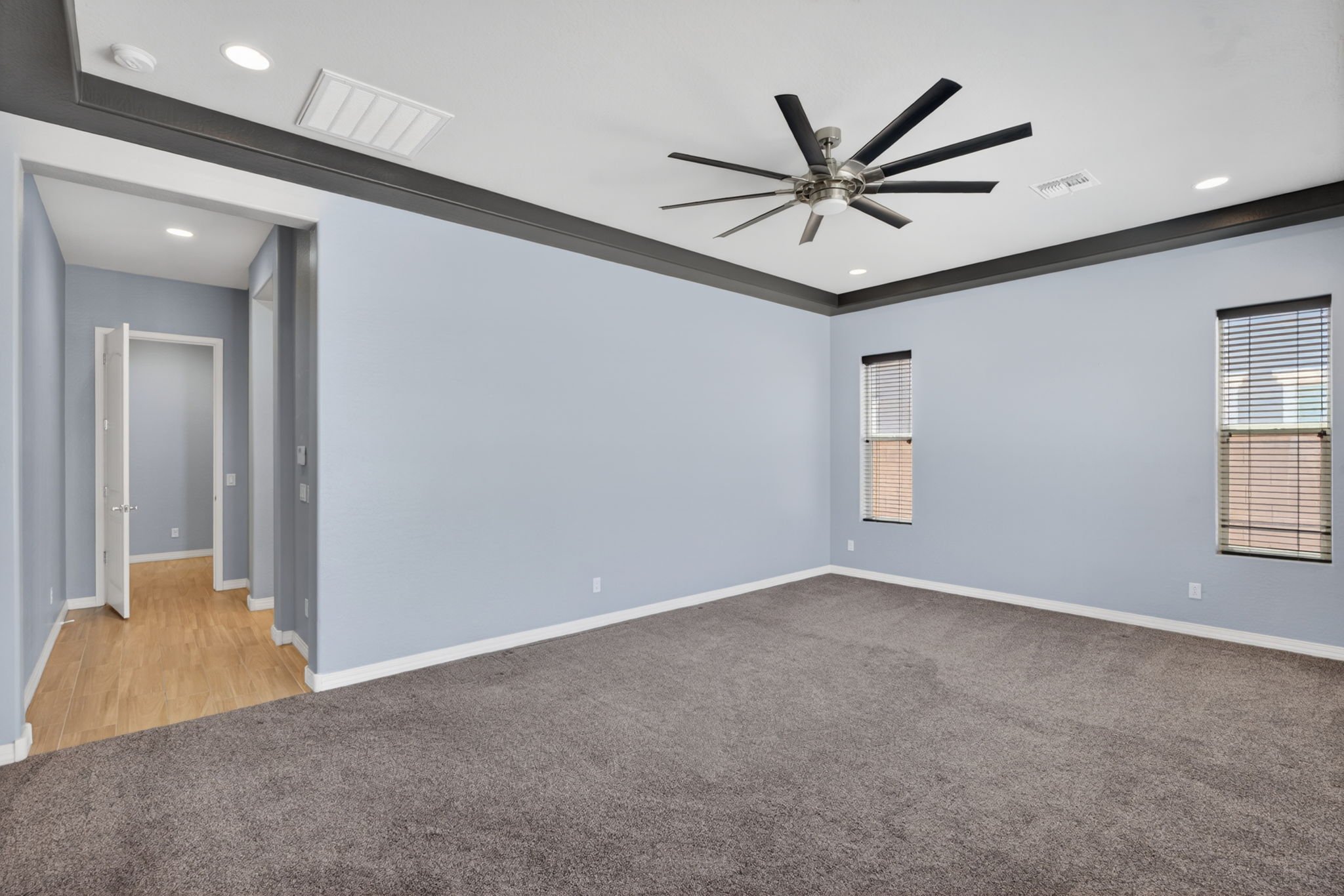 Empty living room with light blue walls, gray carpet, windows with blinds, and a ceiling fan.