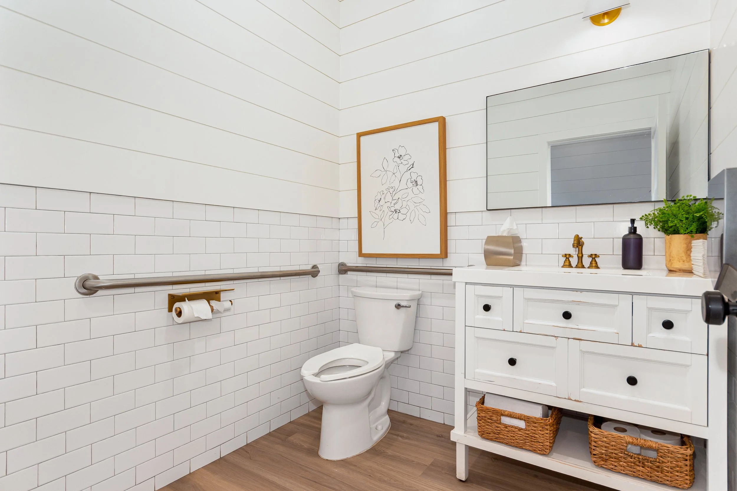 A bathroom with white subway tile walls, a toilet, a white cabinet with black knobs, a framed floral artwork, a large mirror, a potted green plant, a purple soap dispenser, and wicker baskets underneath the cabinet.