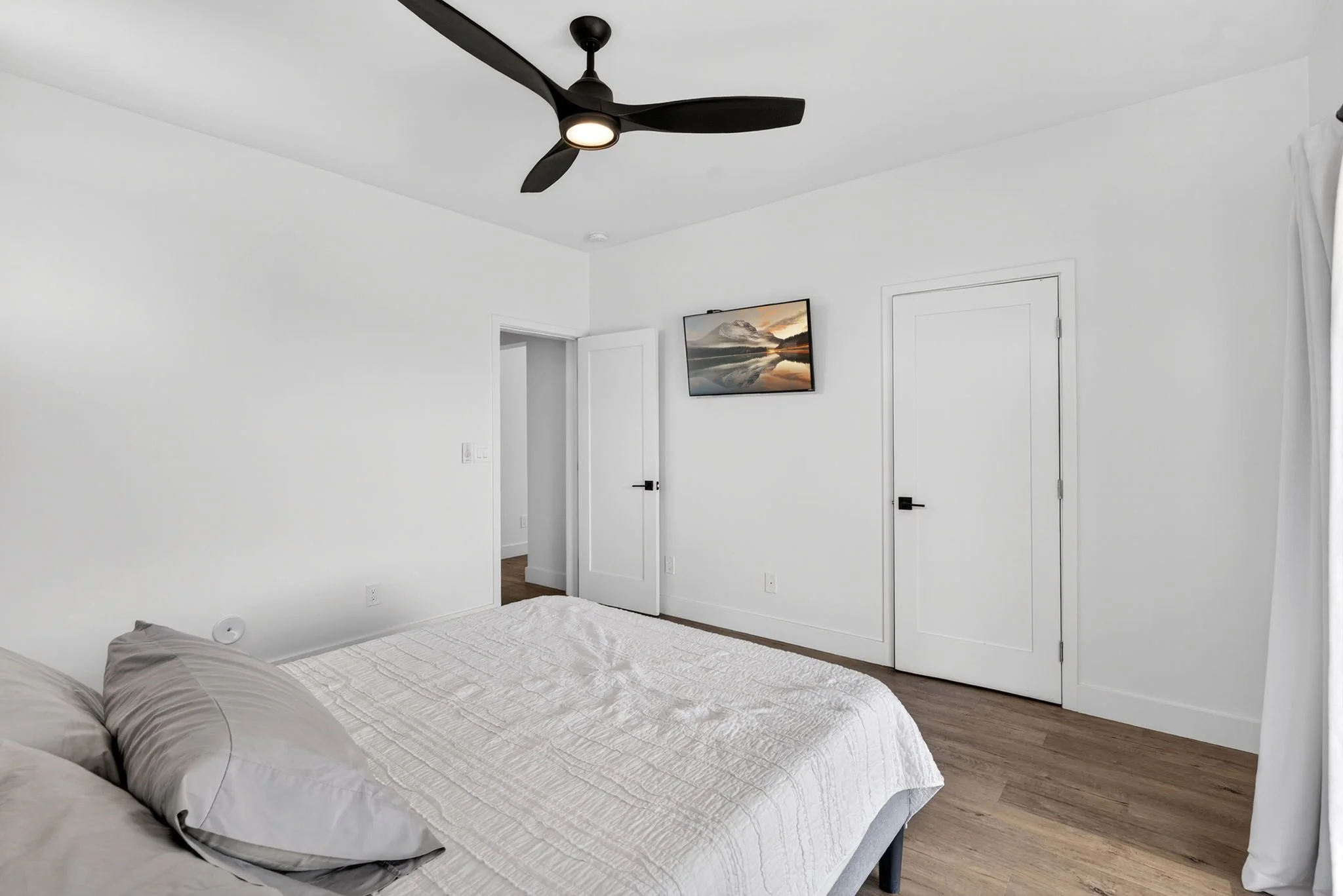 A bedroom with a bed covered in a white quilt and two gray pillows, wood flooring, white walls, a flush door, a wall-mounted TV showing a mountain reflection in water, a ceiling fan with three black blades and a light, and a white curtain on the righ