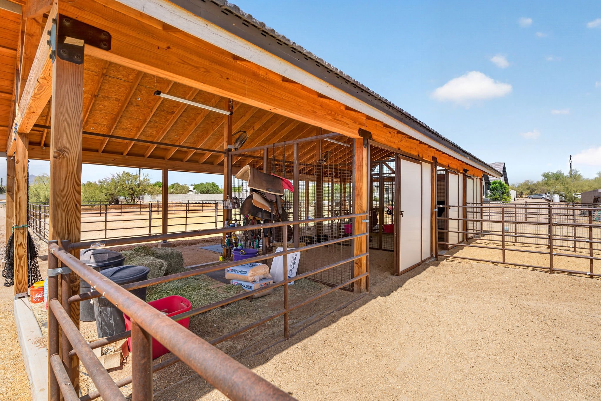 View of a wooden outdoor horse stable with turn-out area under a clear blue sky. The structure features a pitched roof, metal fencing, and a partitioned section for animals. Various equipment and supplies are attached or stored around the stable.