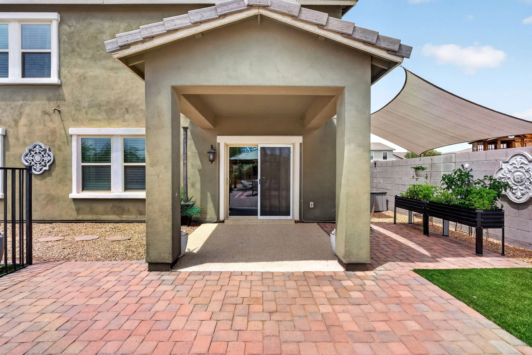 Backyard patio with paver stone flooring, a small covered area with a roof, potted plants, a bench, and decorative wall accents. There's a fence and a shade sail on the right side.