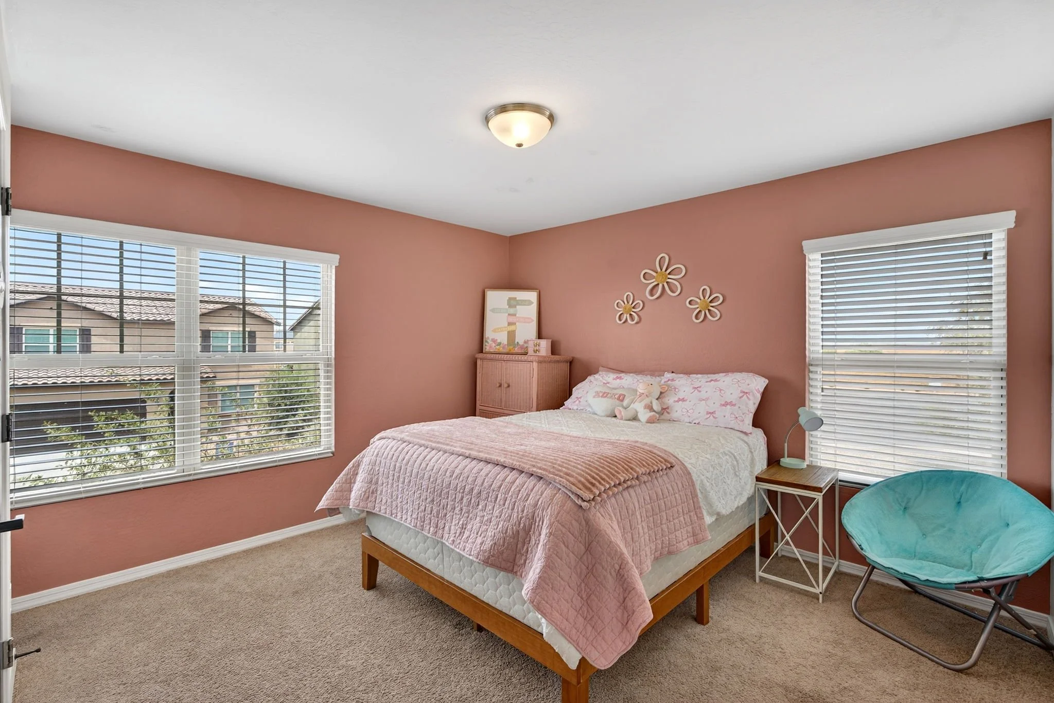 A bedroom with pink walls, two large windows with white blinds, a bed with a pink quilt and stuffed animal, a small nightstand with a lamp, and a corner chair with teal cushion.