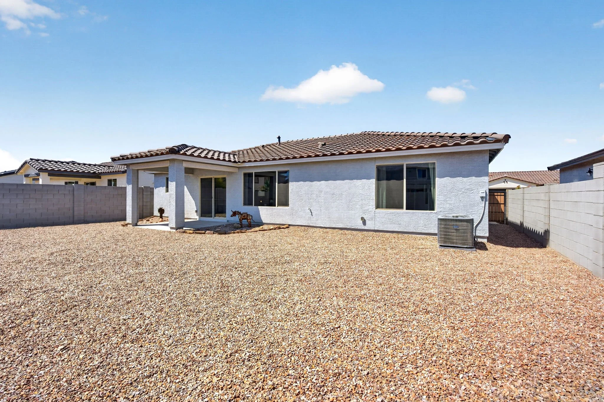 Backyard view of a single-story house with white stucco walls, a tiled roof, and large windows. The yard is covered with gravel, and a cinder block wall surrounds the property. There is an air conditioning unit and a small decorative horse statue nea