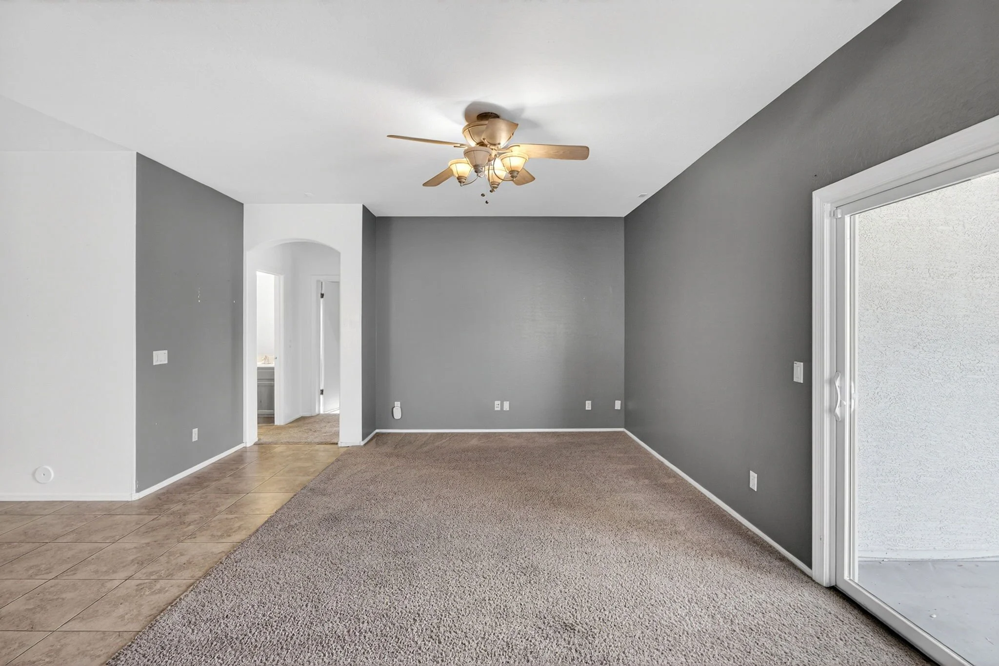Empty living room with gray walls, a ceiling fan with lights, hardwood and carpet flooring, and a sliding glass door.