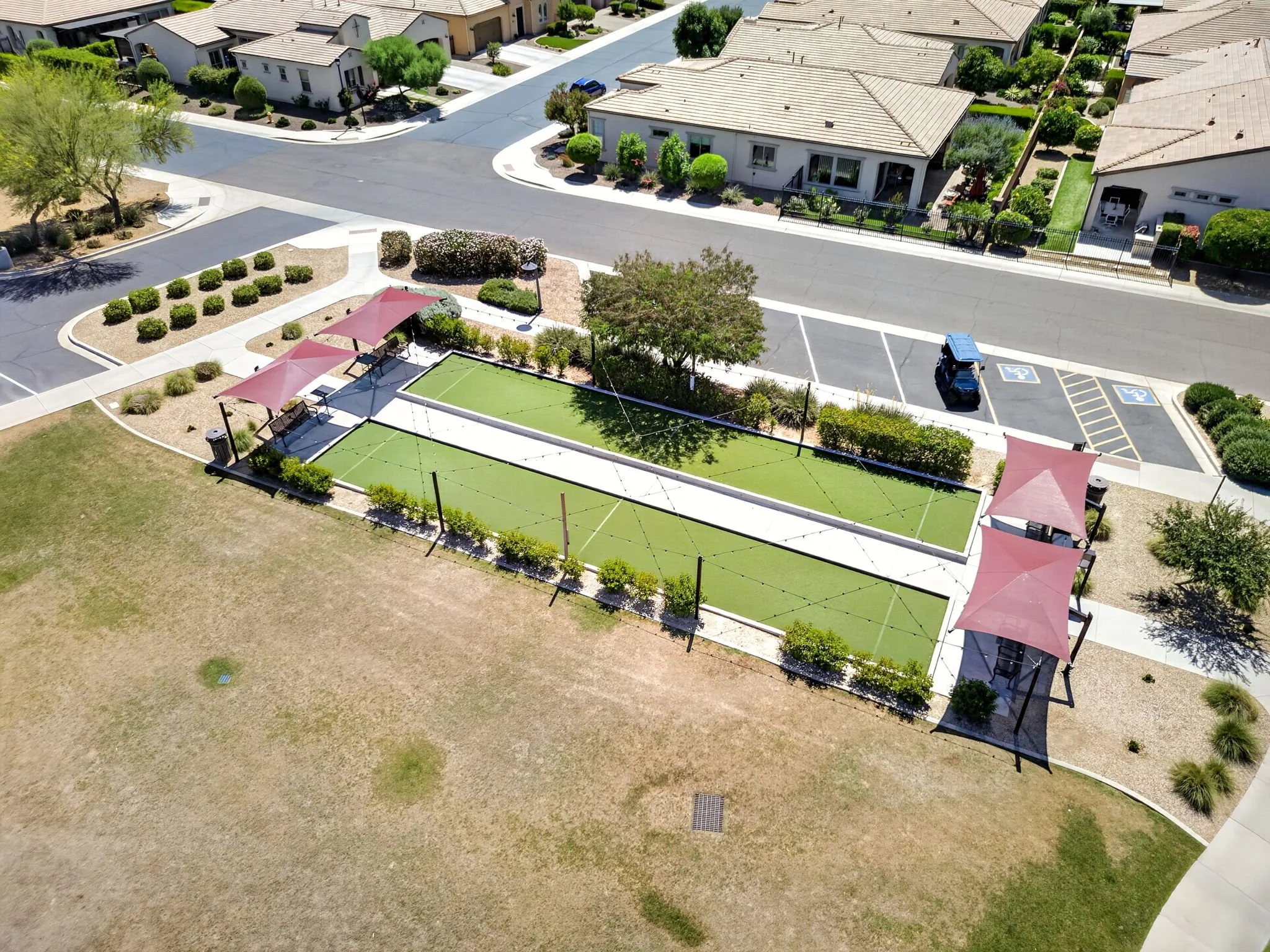 An aerial view of a residential neighborhood featuring houses with tiled roofs, green shrubs, and trees. There is a small recreational area with a shaded seating area, artificial turf, and a playground, along with designated parking spaces including 