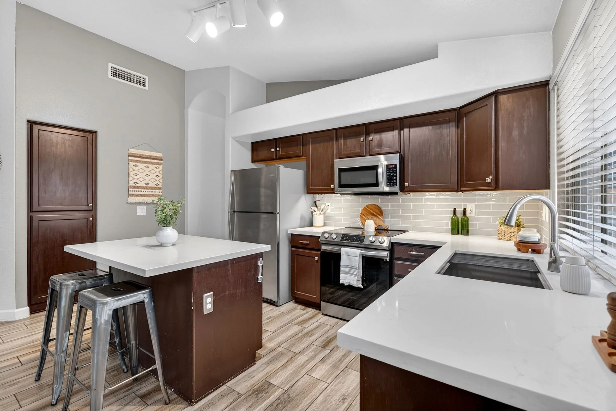 Modern kitchen with wooden cabinets, white countertops, stainless steel refrigerator and microwave, kitchen island with stools, and window with blinds.