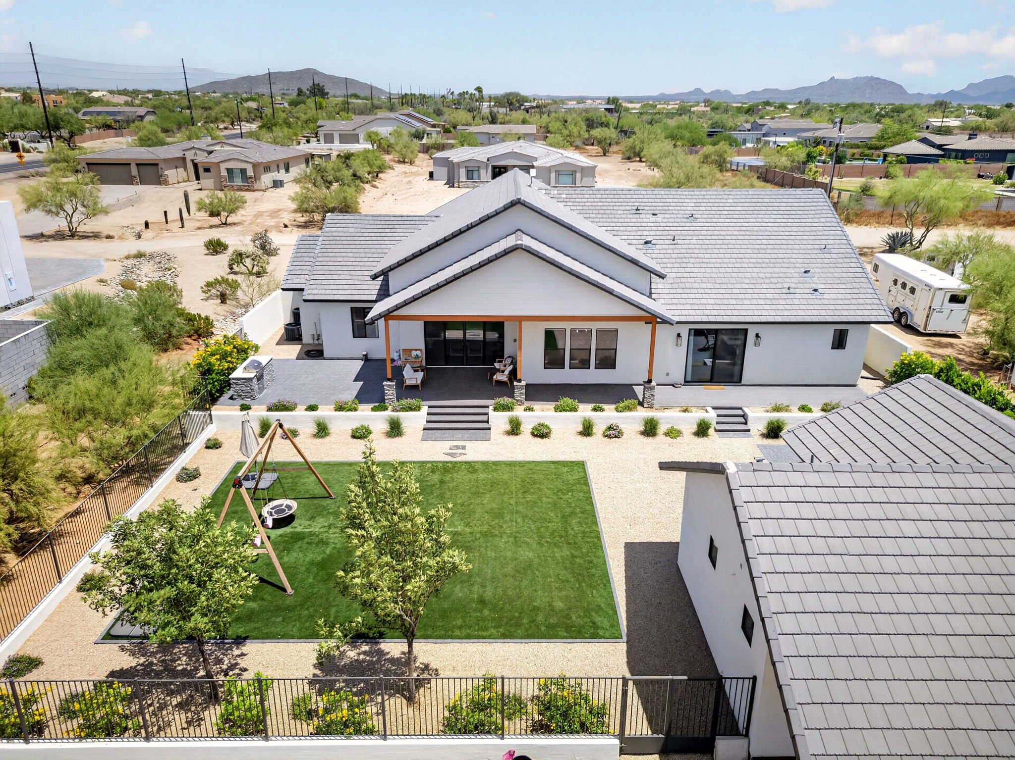 A backyard with a well-maintained lawn, trees, a swing set, and a paved patio area with outdoor furniture. The yard is surrounded by a black metal fence and is adjacent to neighboring houses with desert landscaping and mountainous terrain in the back