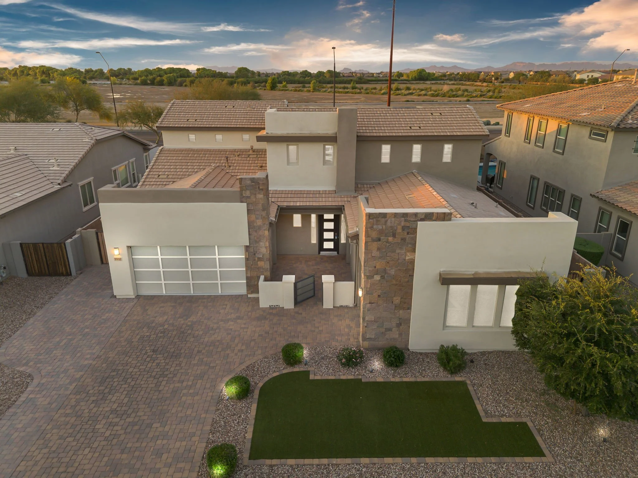 Aerial view of a modern house with a paved driveway, a small lawn, and neighboring houses under a clear sky at sunset.