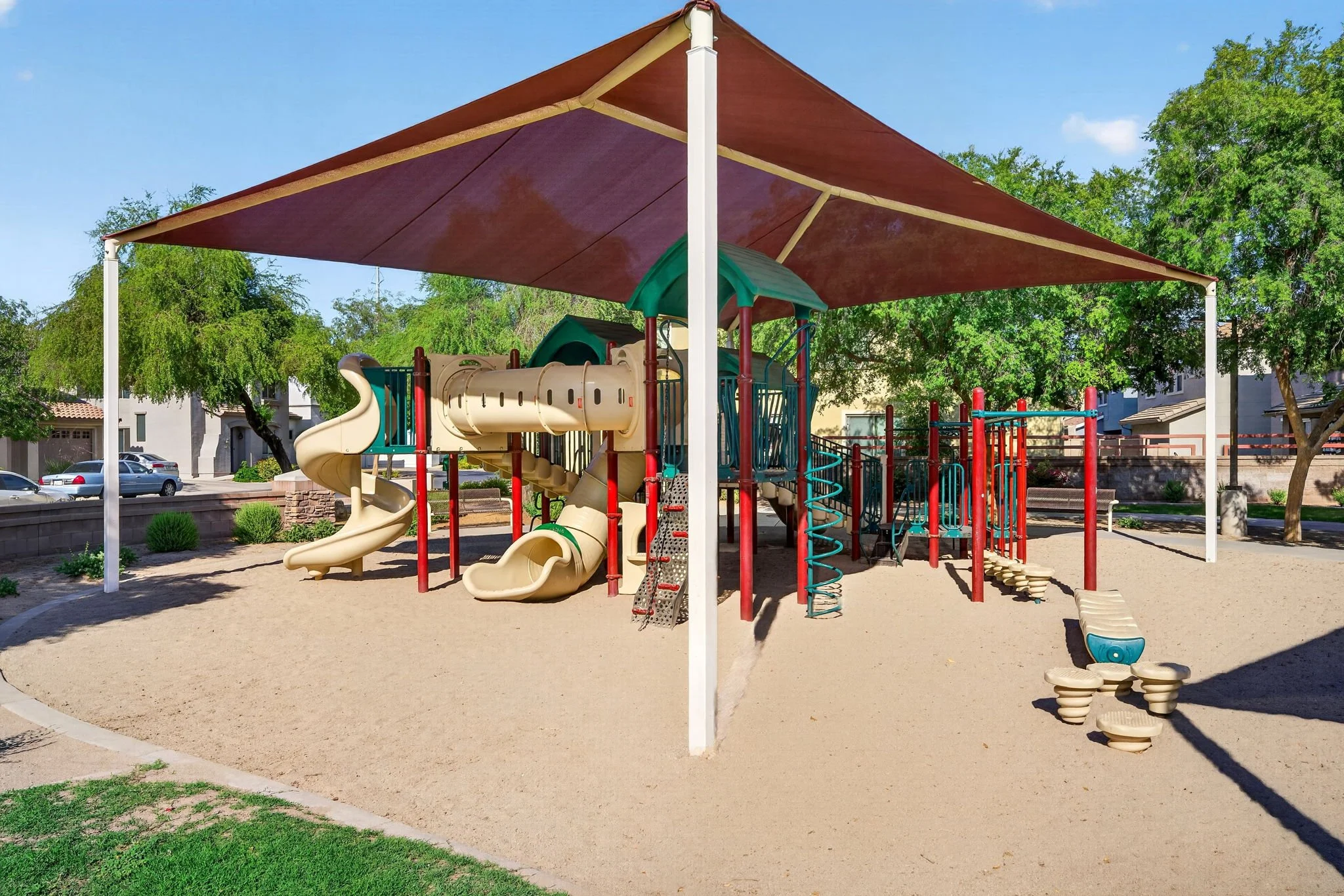 A colorful playground covered with a large red shade sail, featuring slides, climbing structures, and spring riders, surrounded by trees and residential houses.