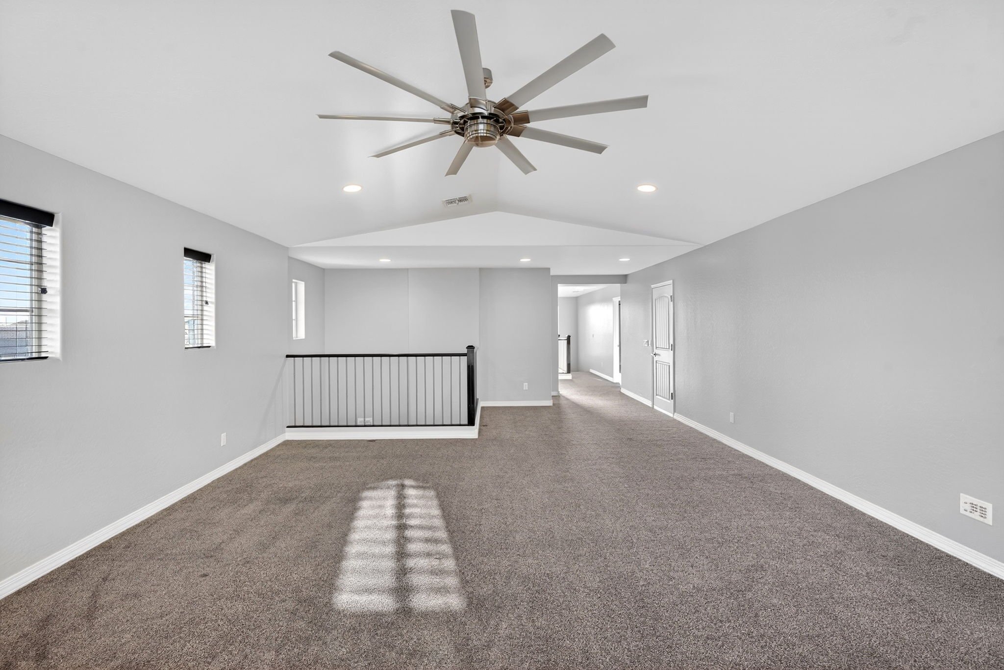 Empty living room with white walls, brown carpet, windows with black blinds, a staircase railing, and ceiling fan.